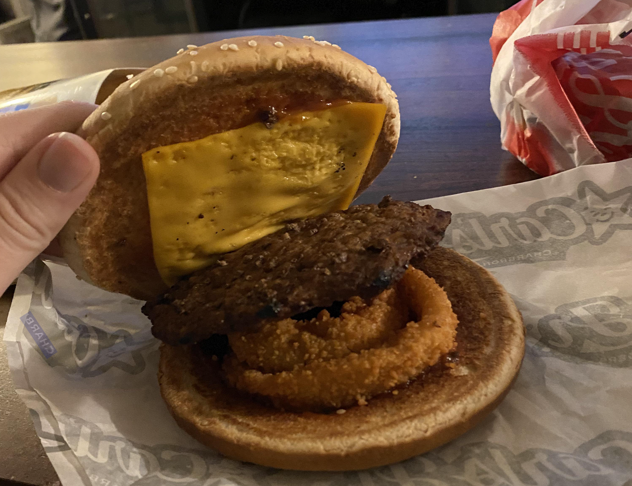 A hand holding open a cheeseburger with a single beef patty and an onion ring on a sesame seed bun.
