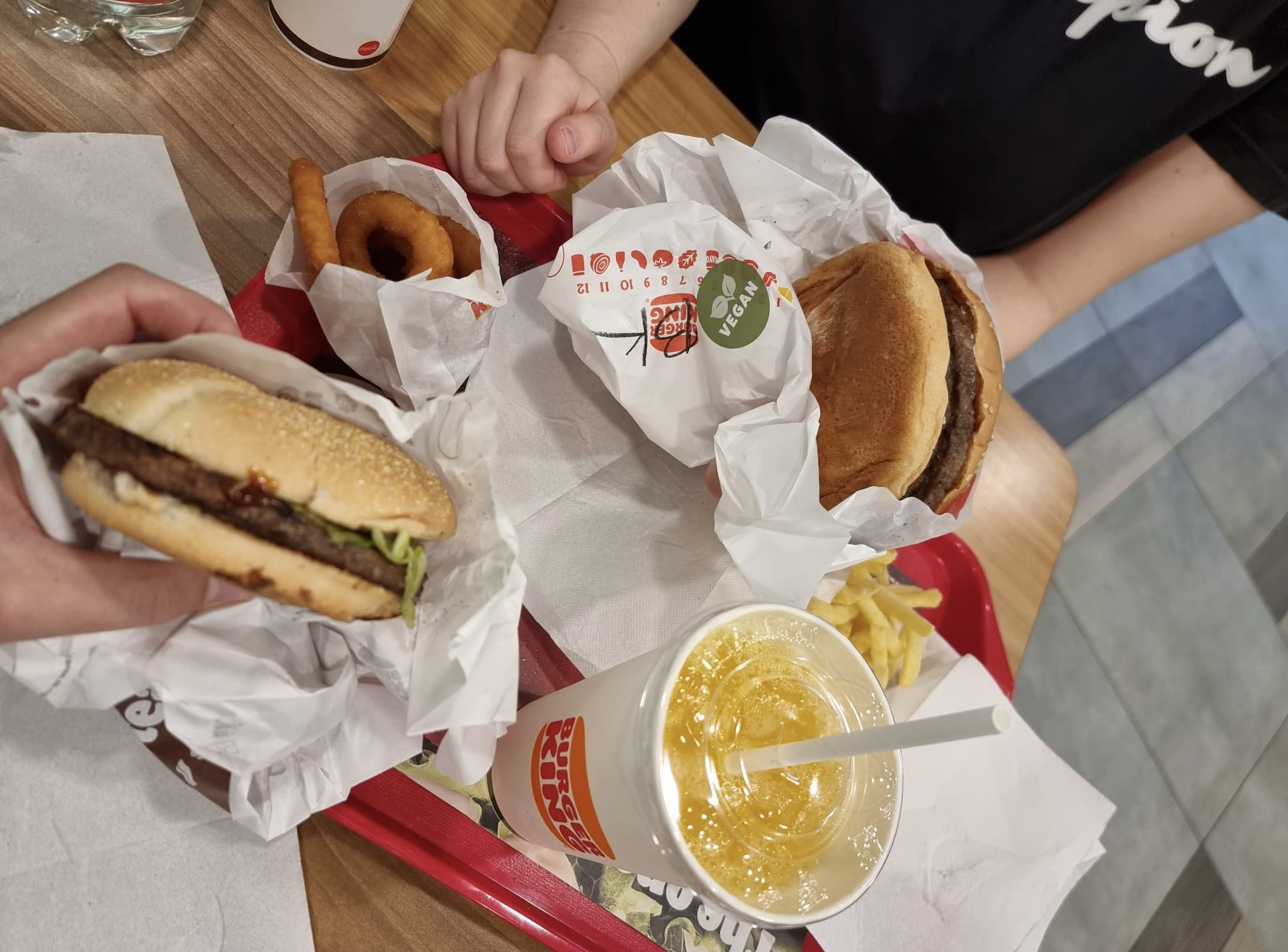 A person holding a burger on a tray with a second burger, onion rings, french fries, and a cup of orange soda.