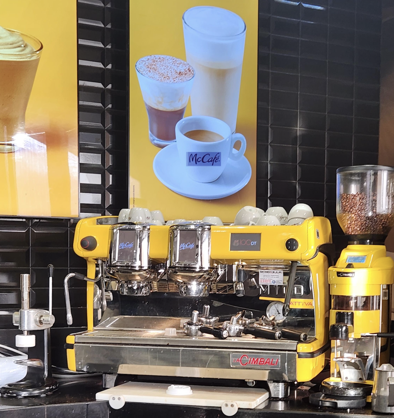 A bright yellow La Cimbali commercial espresso machine with a coffee bean grinder, set against a yellow wall menu featuring pictures of McCafe coffee drinks.