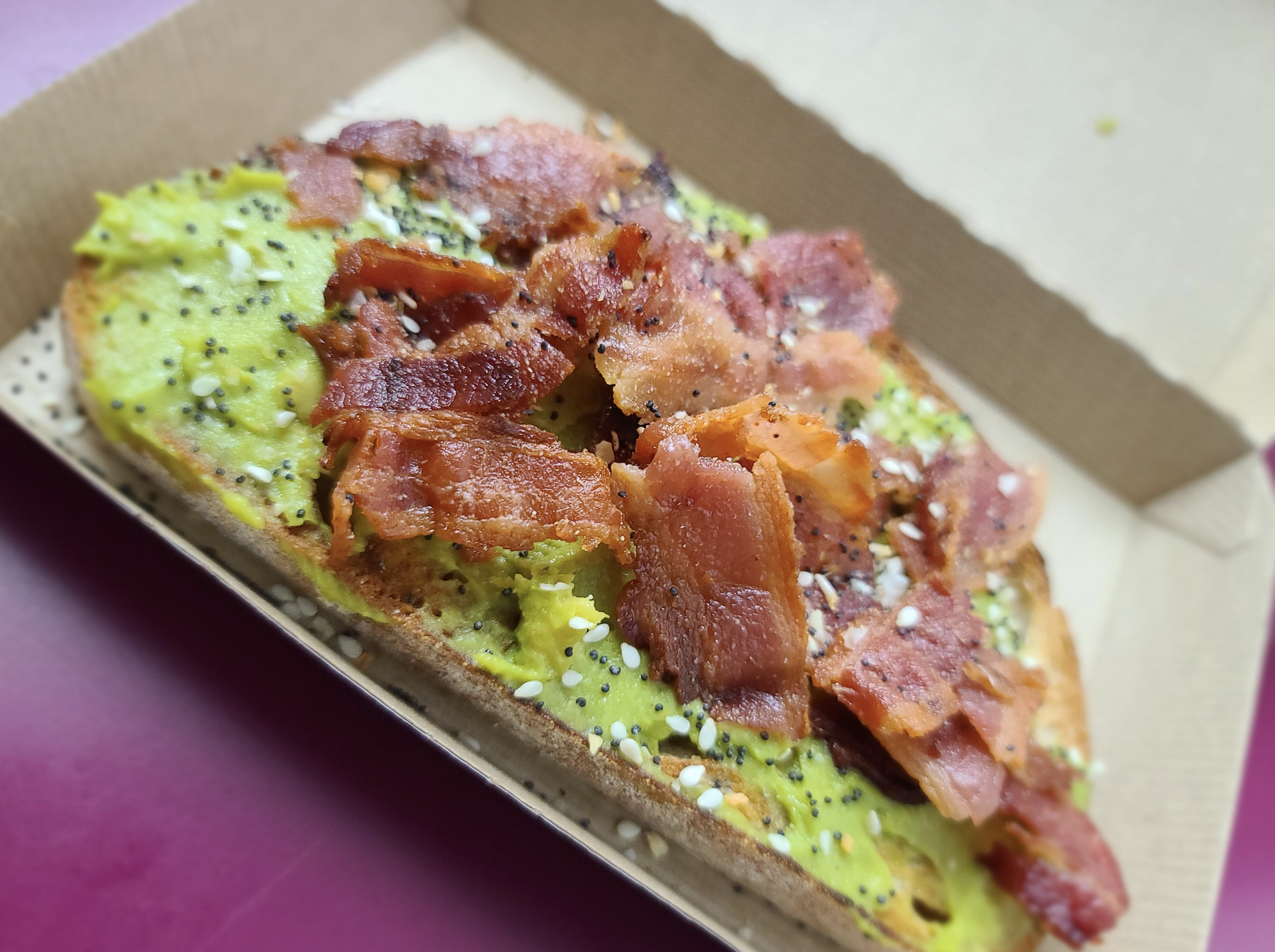 Avocado toast topped with crispy bacon pieces and everything bagel seasoning in a cardboard takeaway container.