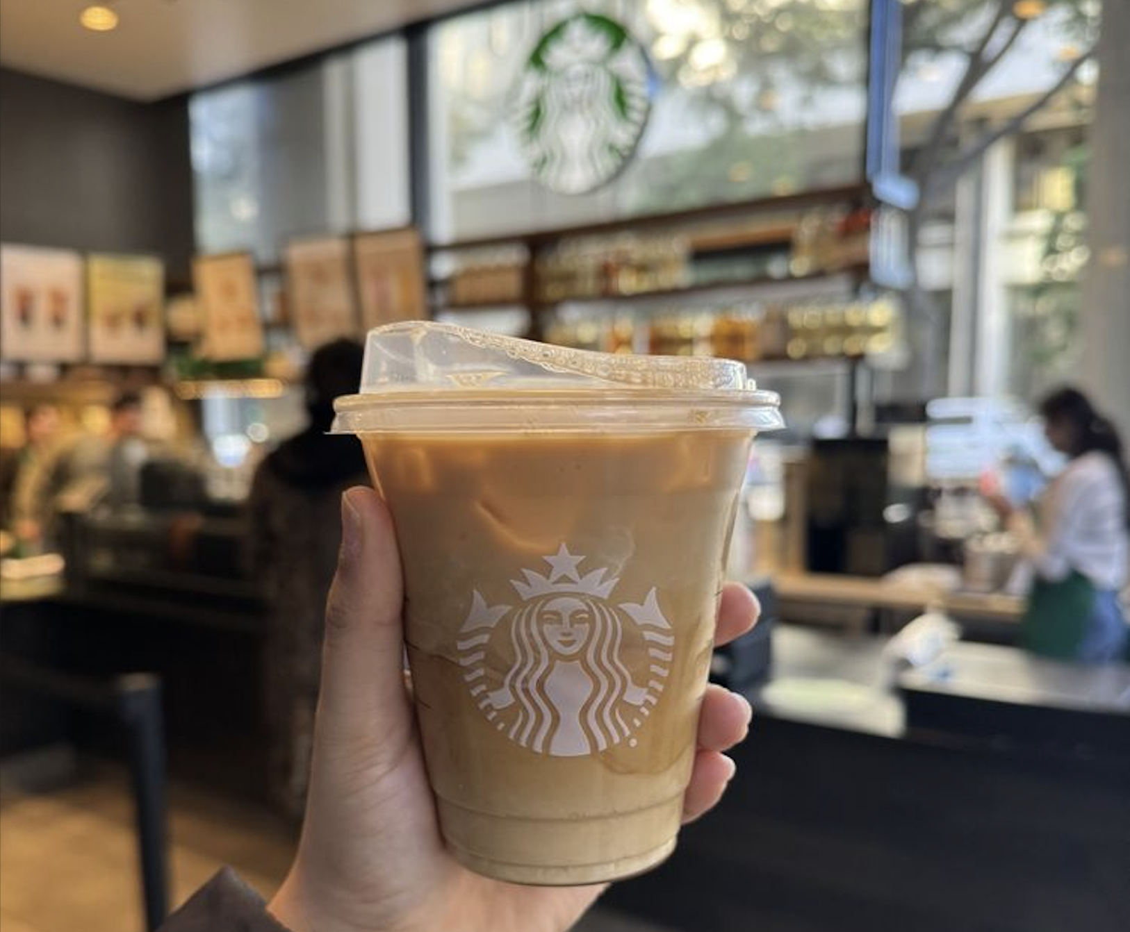 A hand holds an iced Starbucks coffee in a plastic cup inside a café.