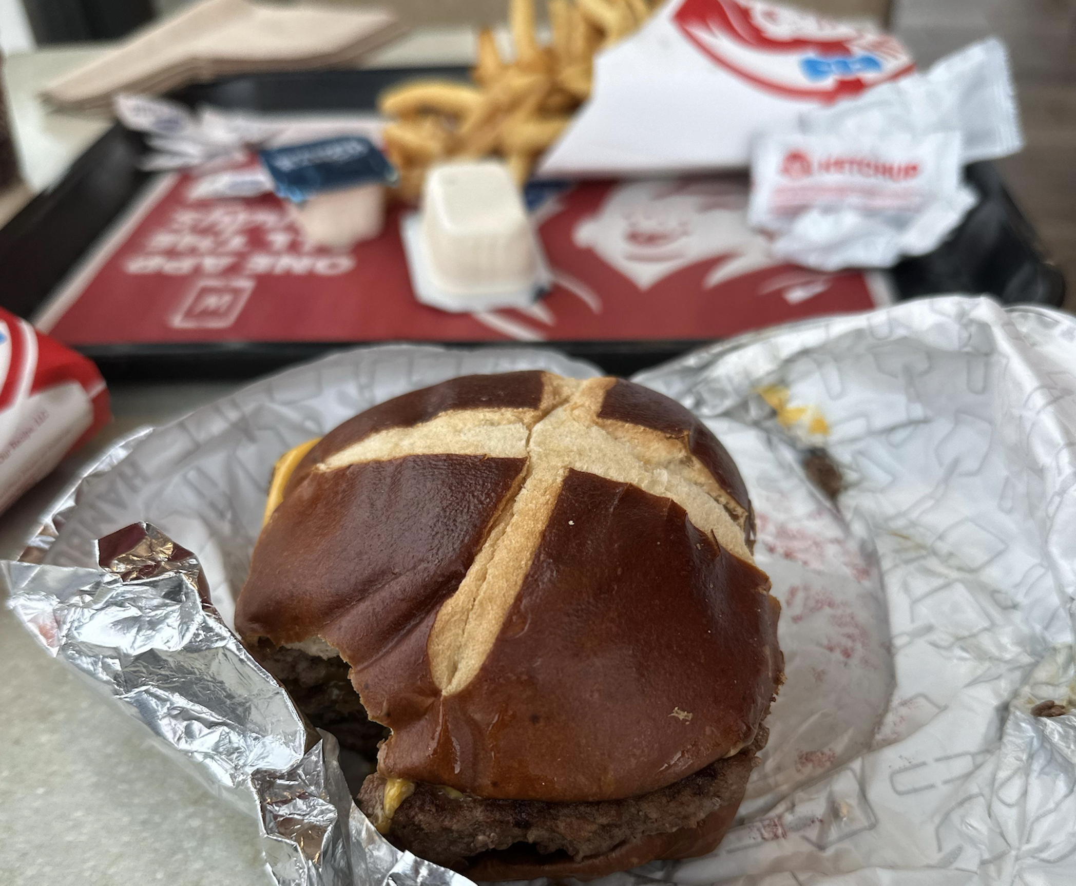 A partially eaten cheeseburger on a pretzel bun in a foil wrapper, with a tray of french fries and condiment packets in the background.