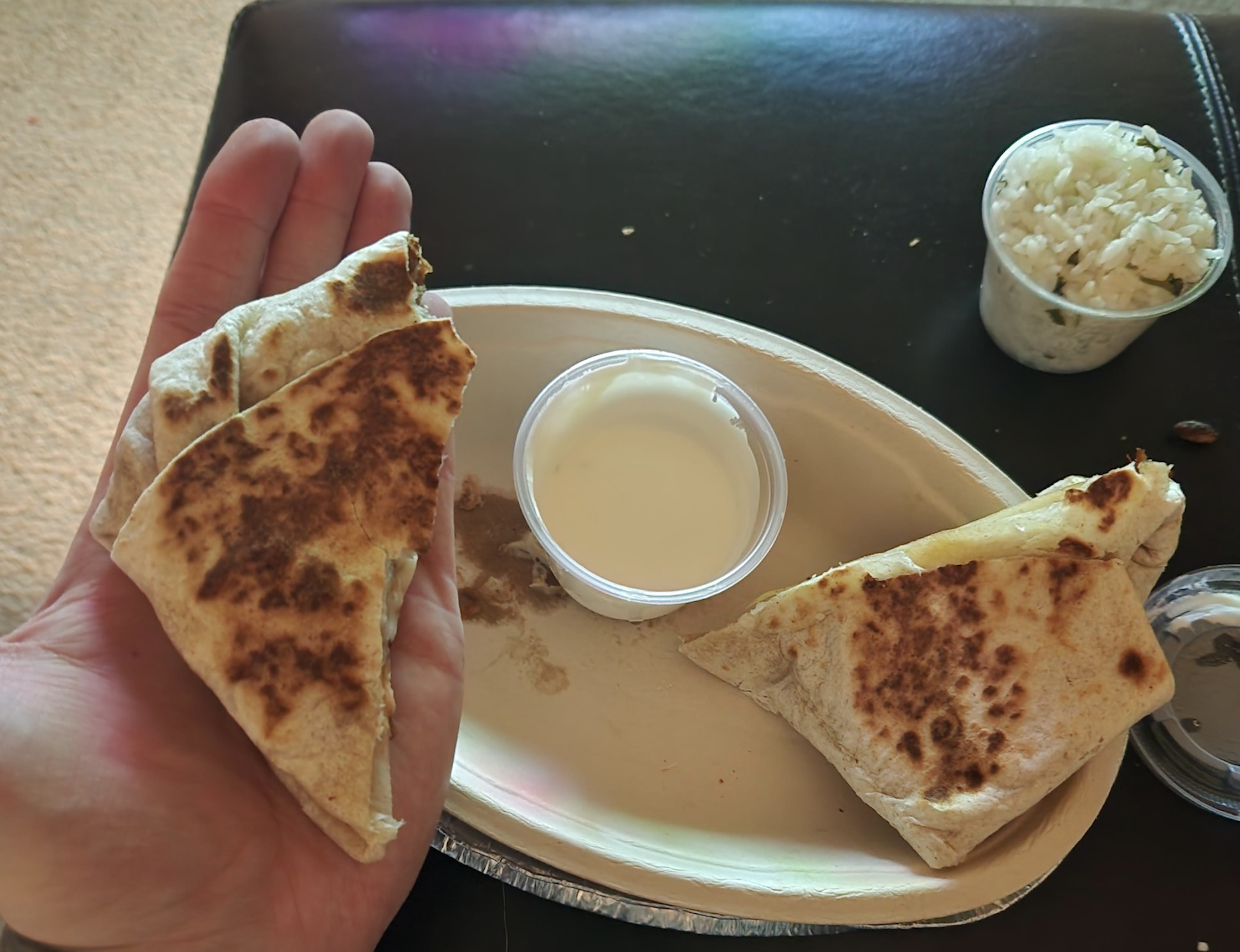 A person holds a toasted tortilla triangle in their hand next to a paper plate with another tortilla triangle, a cup of white sauce, and a small container of rice.