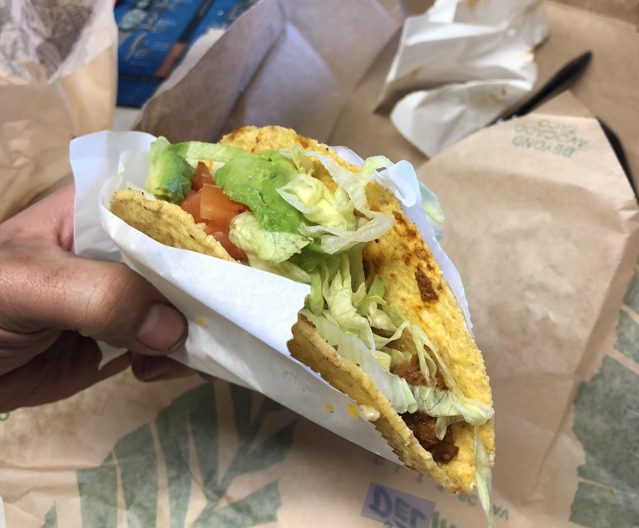 A hand holds a crispy taco in a paper wrapper, filled with ground meat, shredded lettuce, tomato, and avocado.
