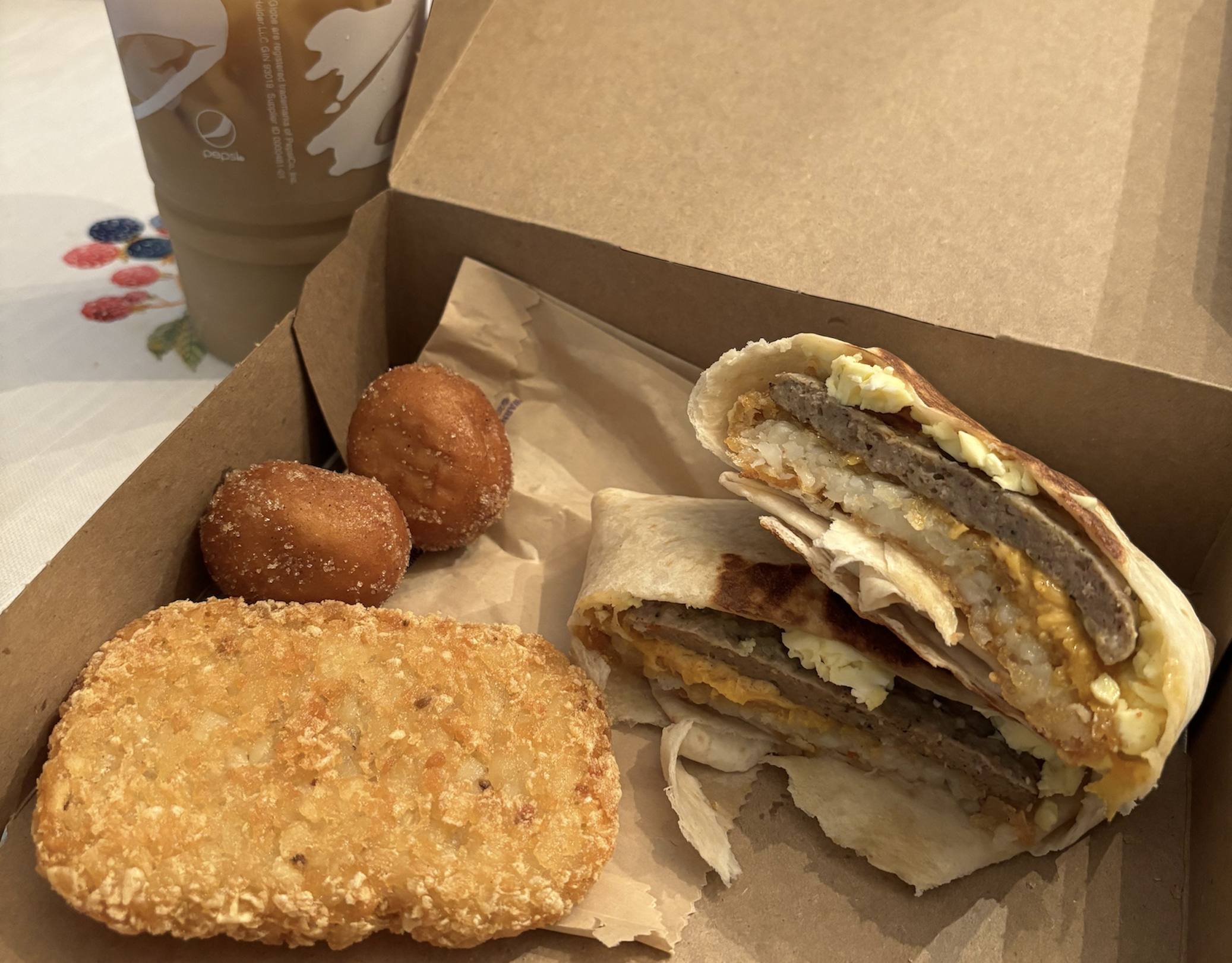 A cardboard takeout box containing a sliced breakfast burrito, a hash brown, and two donut holes, with a Taco Bell drink cup in the background.