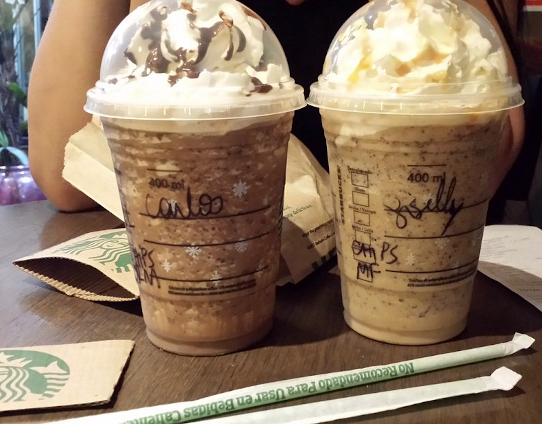 Two Starbucks blended beverages in plastic cups topped with whipped cream and drizzle, with names written on the sides.