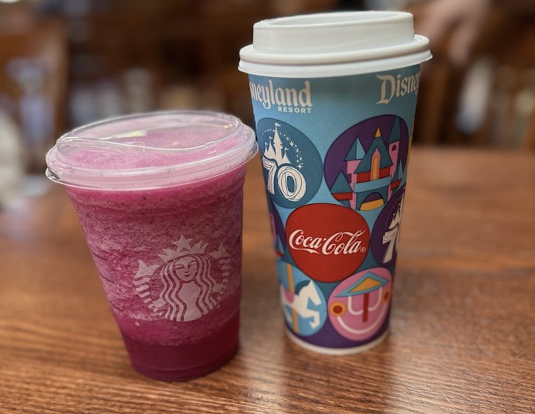A clear plastic cup of pink blended drink with a Starbucks logo next to a paper Disneyland Resort 70th anniversary cup with a Coca-Cola logo.