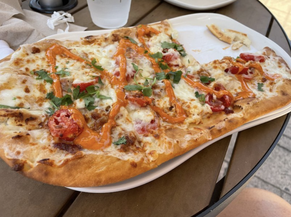 Flatbread pizza, one of Panera's fast-food menus, topped with melted cheese, cherry tomatoes, fresh herbs, and a spicy orange drizzle on a white rectangular plate.