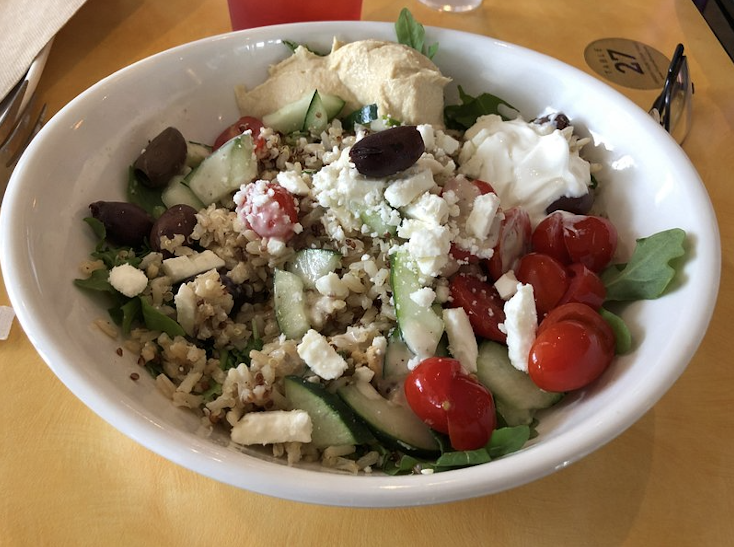 A grain bowl with quinoa, cucumber, cherry tomatoes, olives, feta cheese, hummus, and a dollop of yogurt in a white bowl.
