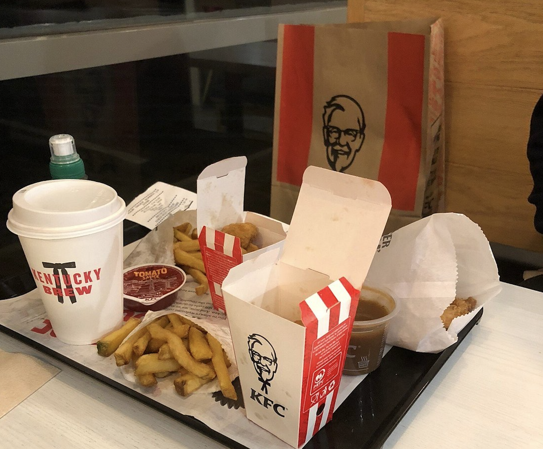 A tray of KFC fast food including two open takeout boxes, French fries, a cup of Kentucky Brew coffee, a container of gravy, and a ketchup packet next to a KFC paper bag.