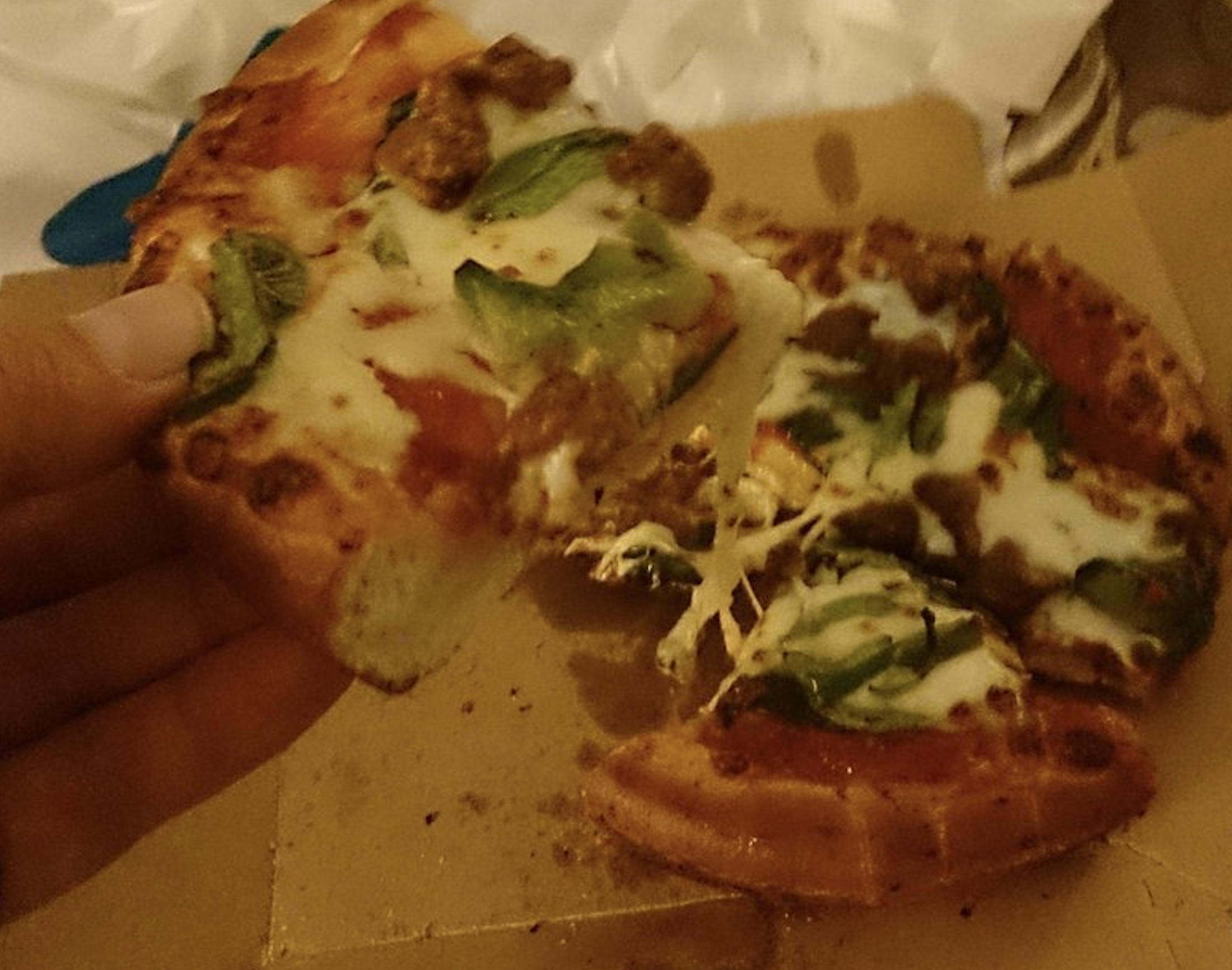 A hand holding a slice of sausage and green pepper pizza with melting cheese.