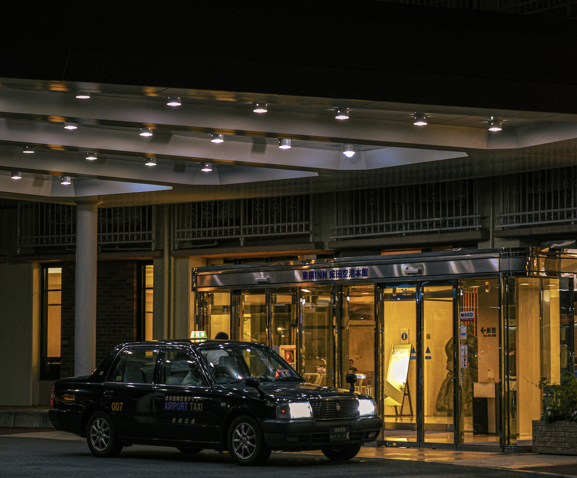 A black airport taxi idling outside the glass entrance of a hotel at night.