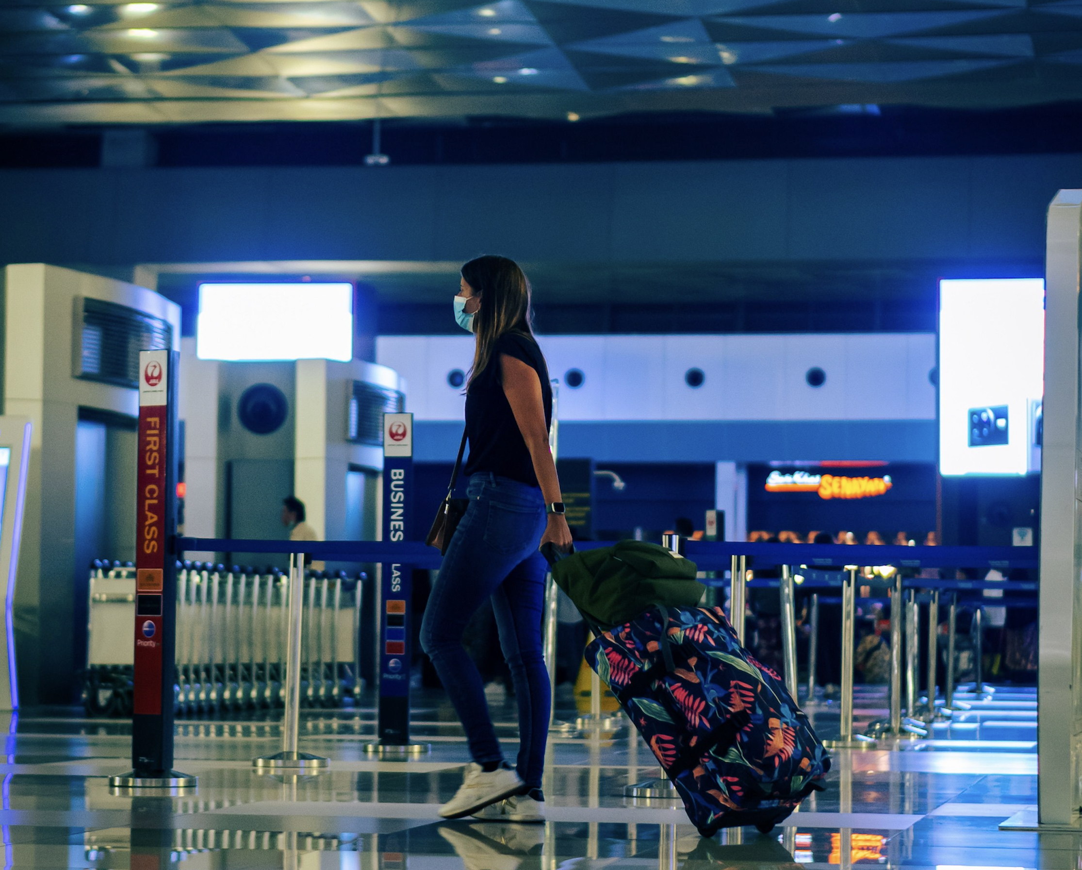 A woman wearing a face mask walks through an airport terminal pulling a patterned rolling suitcase.