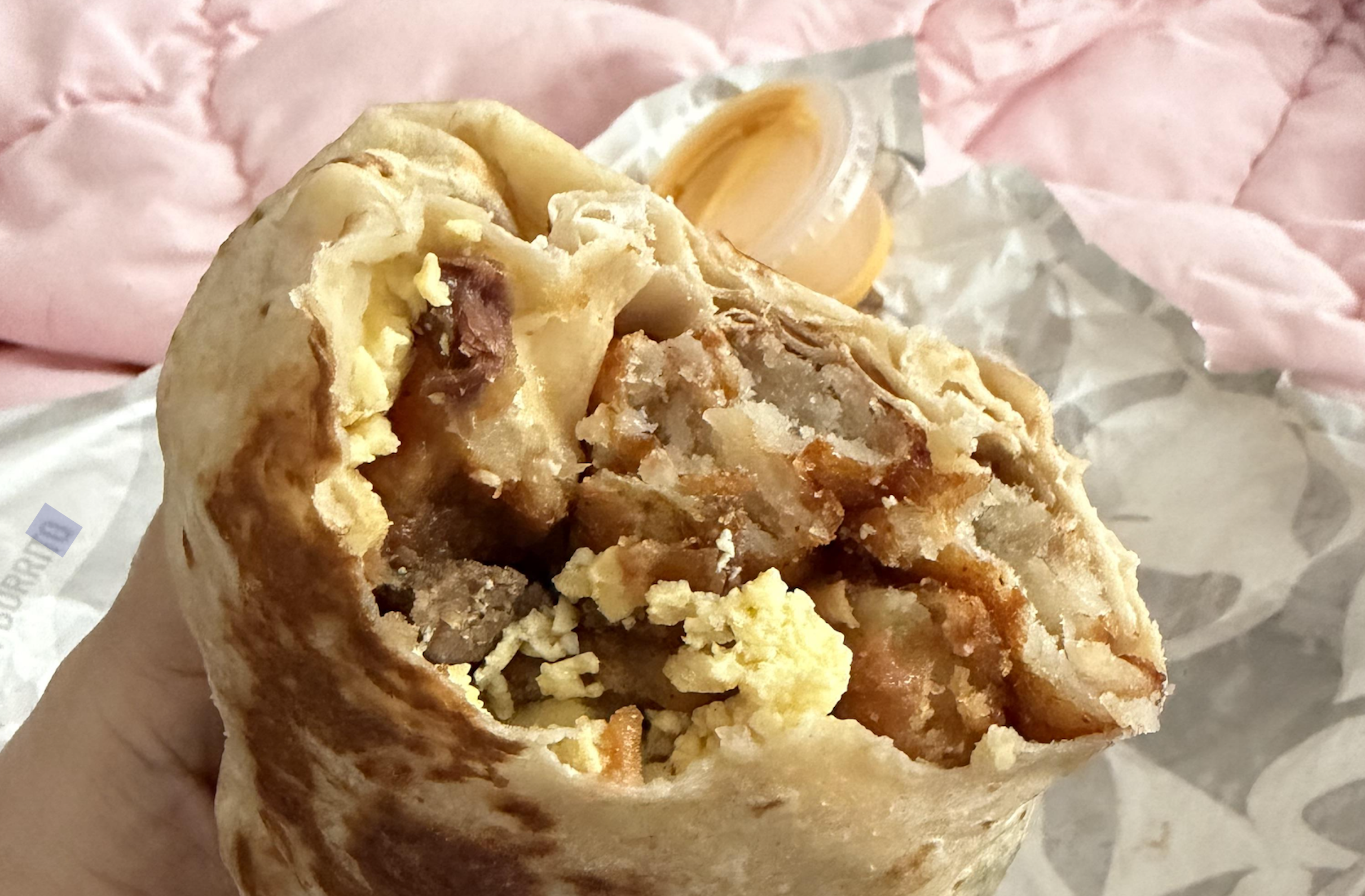 A partially eaten breakfast burrito filled with scrambled eggs, meat, and fried potatoes, with a small container of sauce in the background.