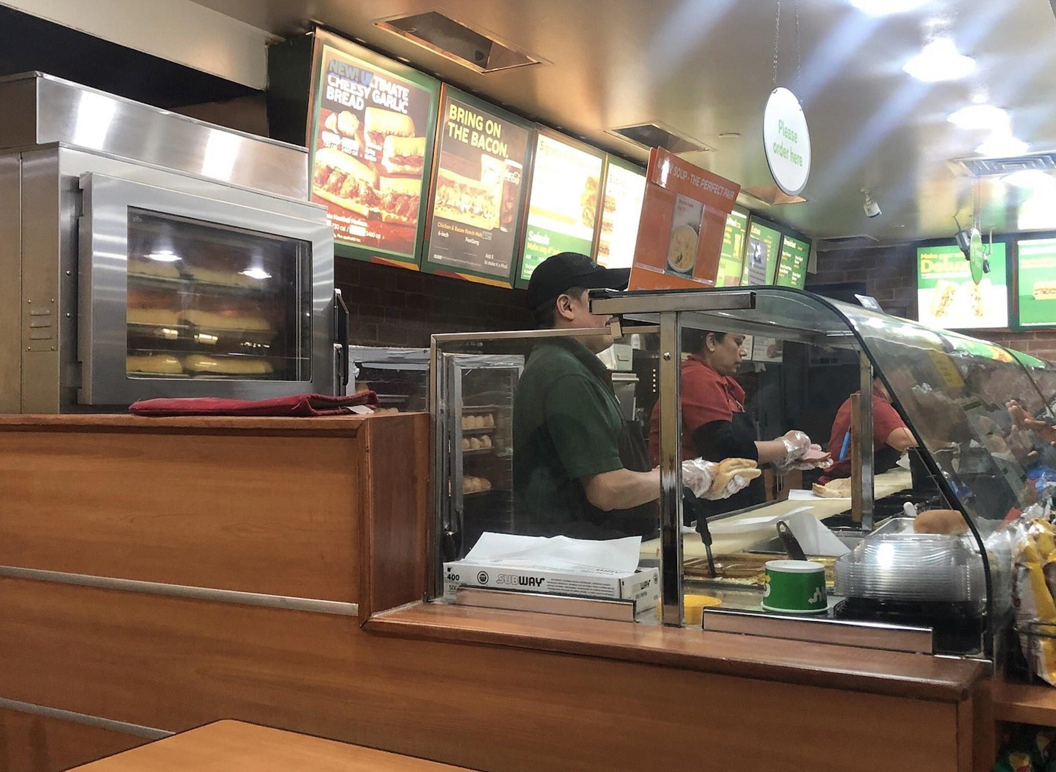 Two Subway employees in uniform prepare sandwiches behind a glass food counter, with menu boards displayed on the wall behind them.