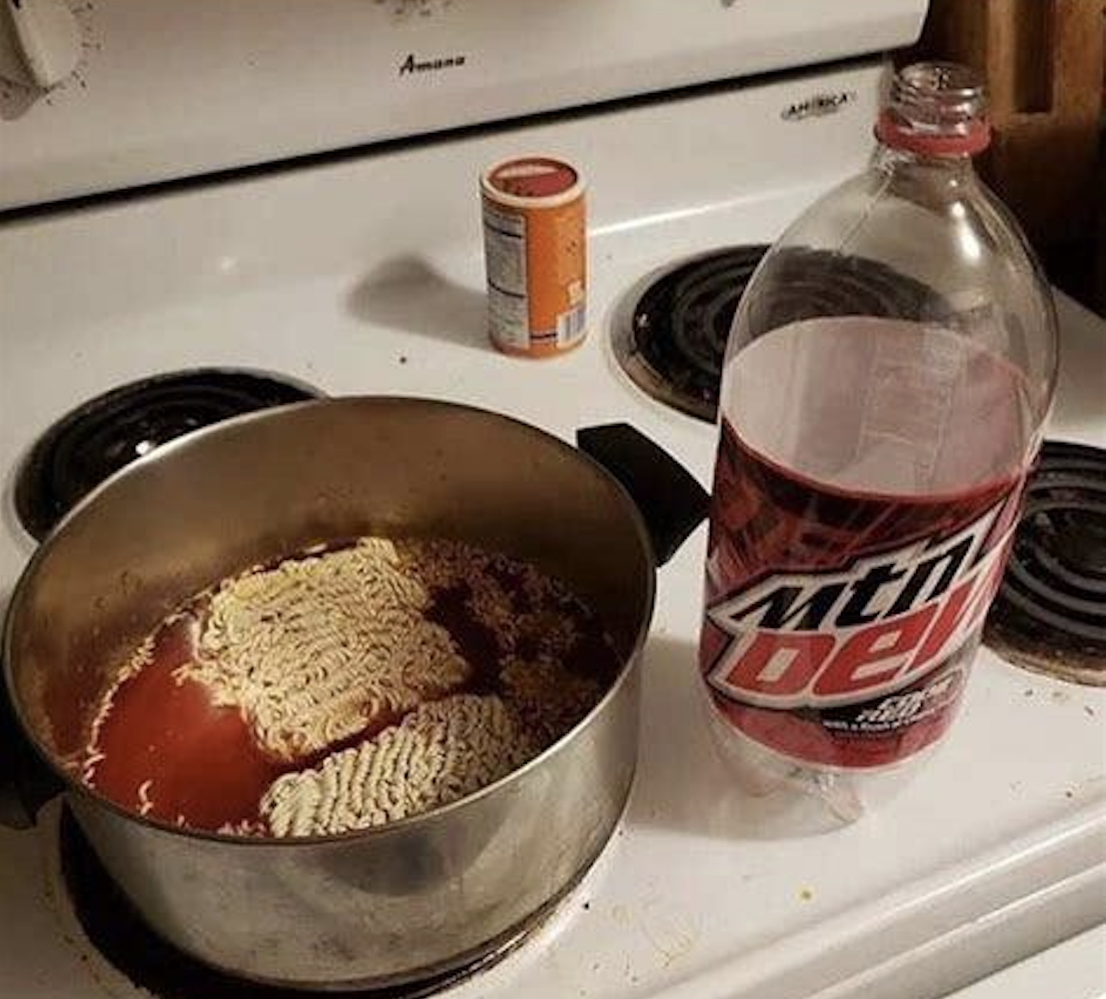 A metal pot containing ramen noodles submerged in red liquid, sitting on a stove next to a bottle of Mountain Dew.