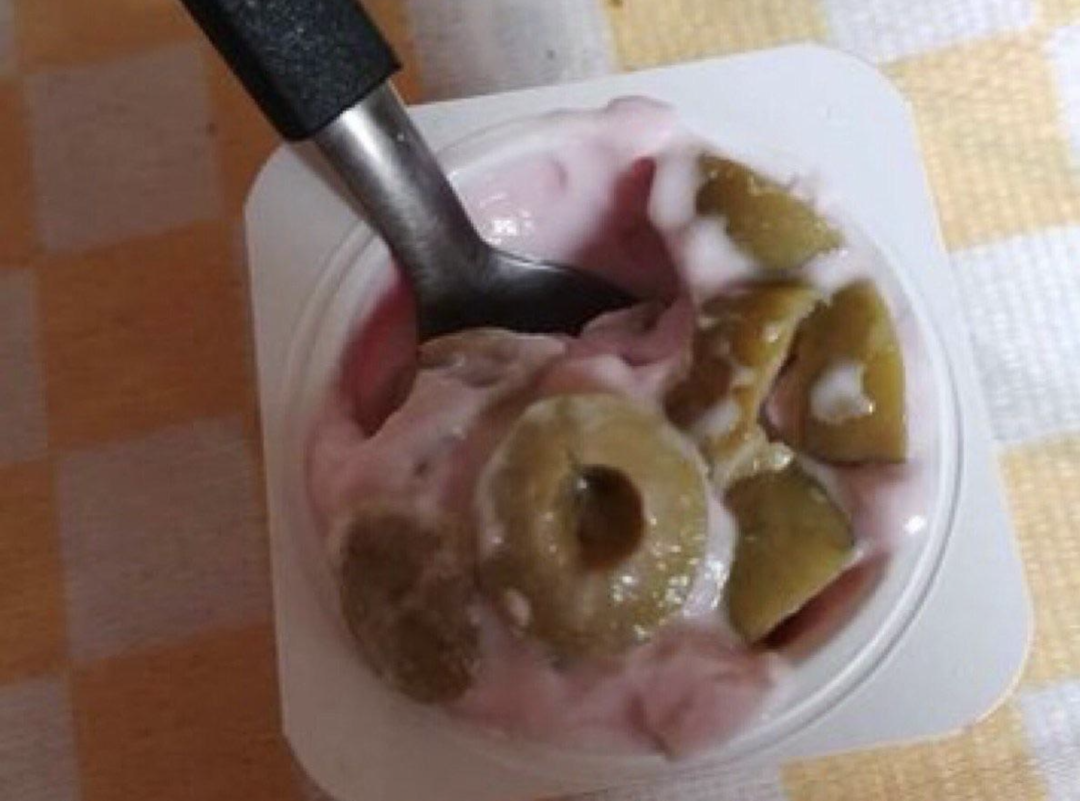 A metal spoon stirs sliced green olives into a cup of pink yogurt.