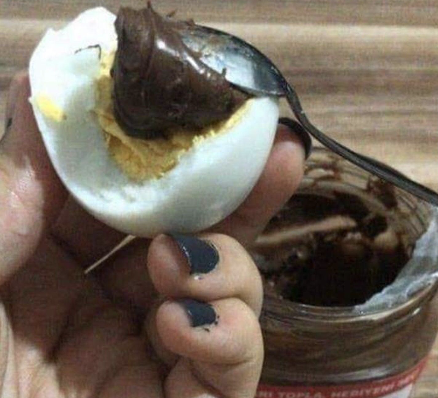 A hand holds a hard-boiled egg being topped with a spoonful of chocolate spread from a jar.
