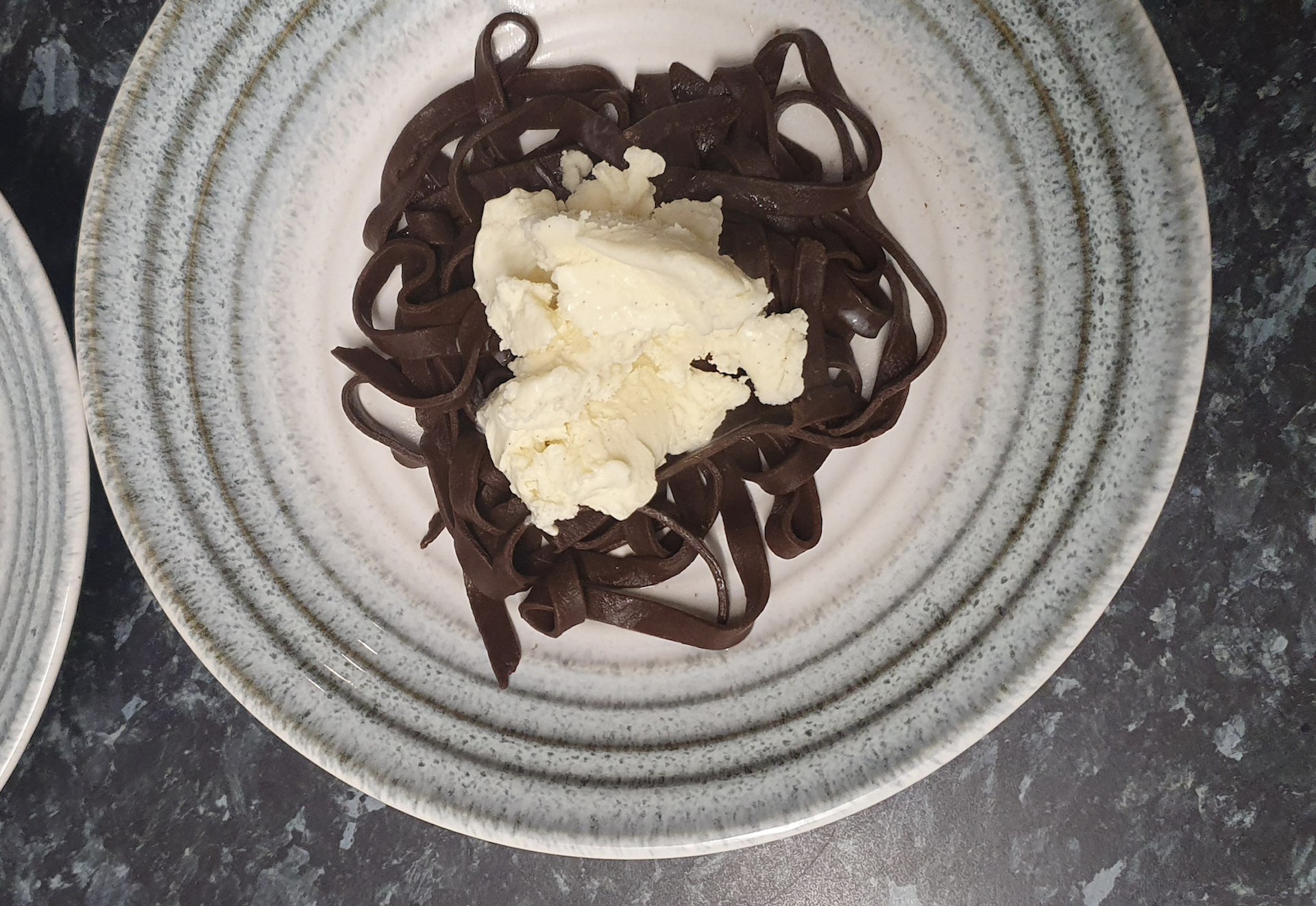 A serving of dark chocolate pasta topped with a scoop of vanilla ice cream on a speckled ceramic plate.