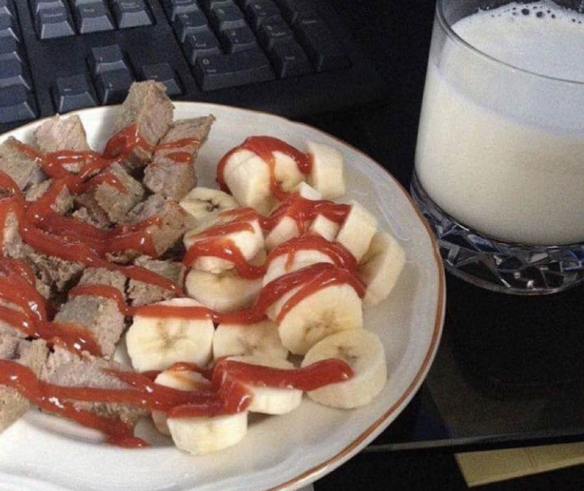 A plate of sliced steak and banana rounds, both drizzled with ketchup, served next to a glass of milk near a computer keyboard.