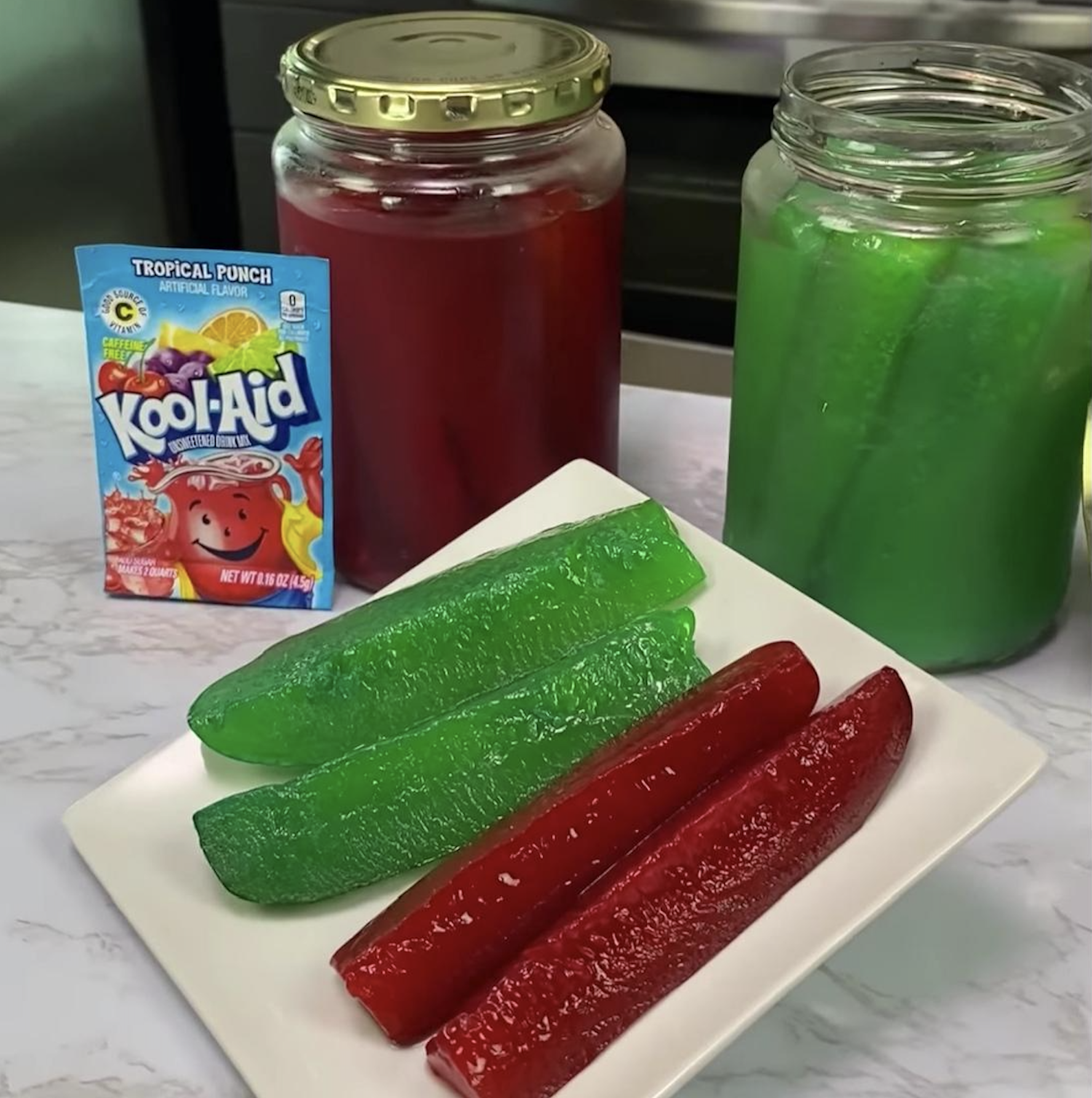 Two bright green and two deep red Kool-Aid pickles on a white plate, with jars of red and green liquid and a Tropical Punch Kool-Aid packet in the background.