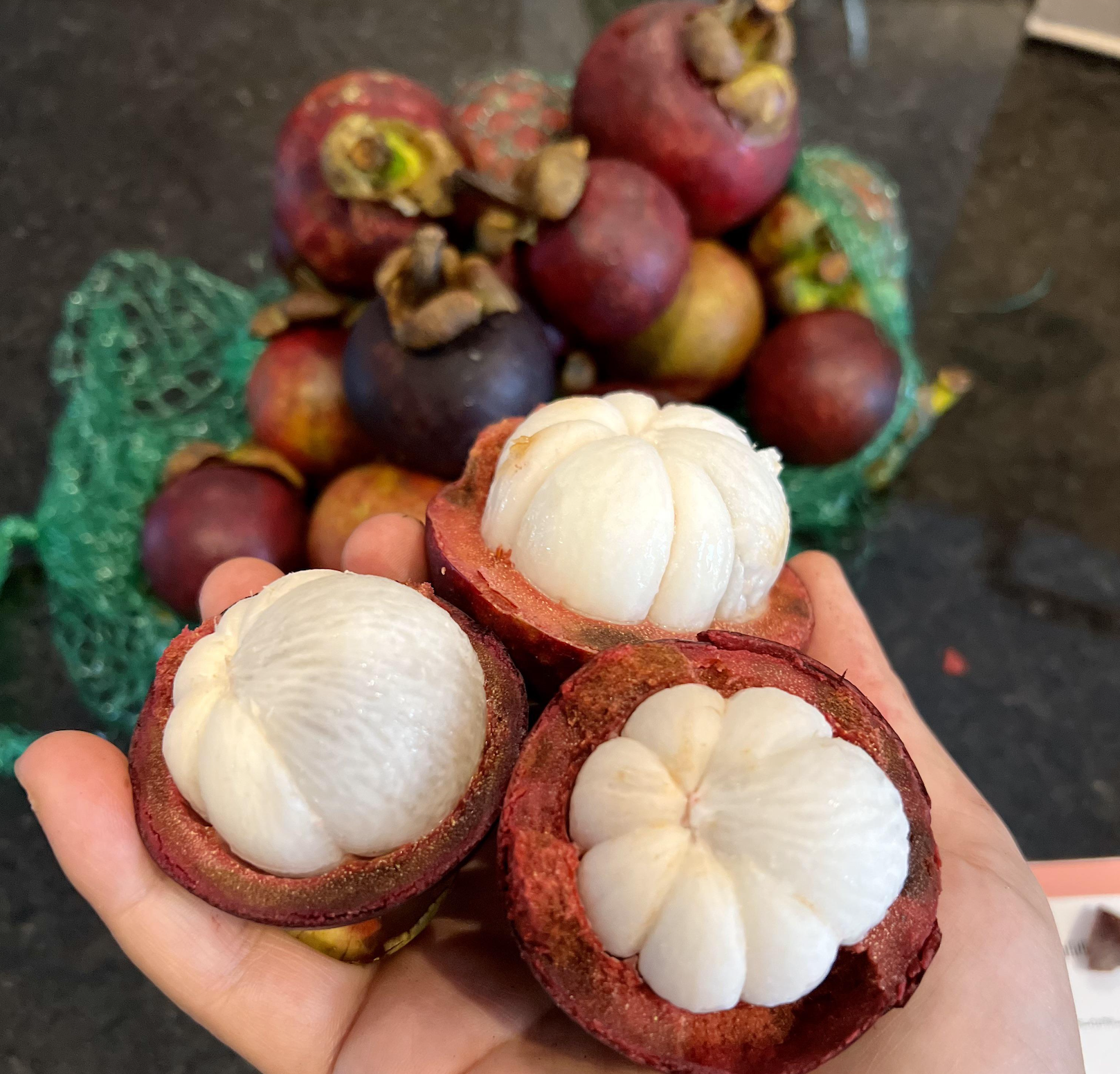 A person’s hand holding three halved mangosteen fruits, revealing their white, segmented flesh, with a pile of whole mangosteens in a green mesh bag in the background.