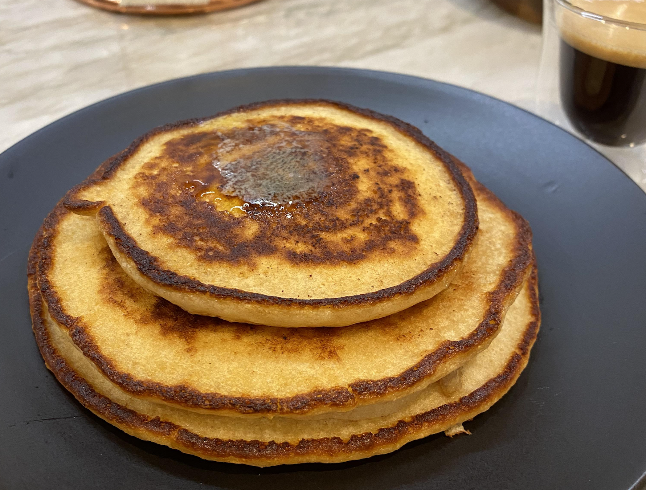A stack of three golden-brown pancakes with a pat of melting butter on top, served on a black plate next to a glass of espresso.