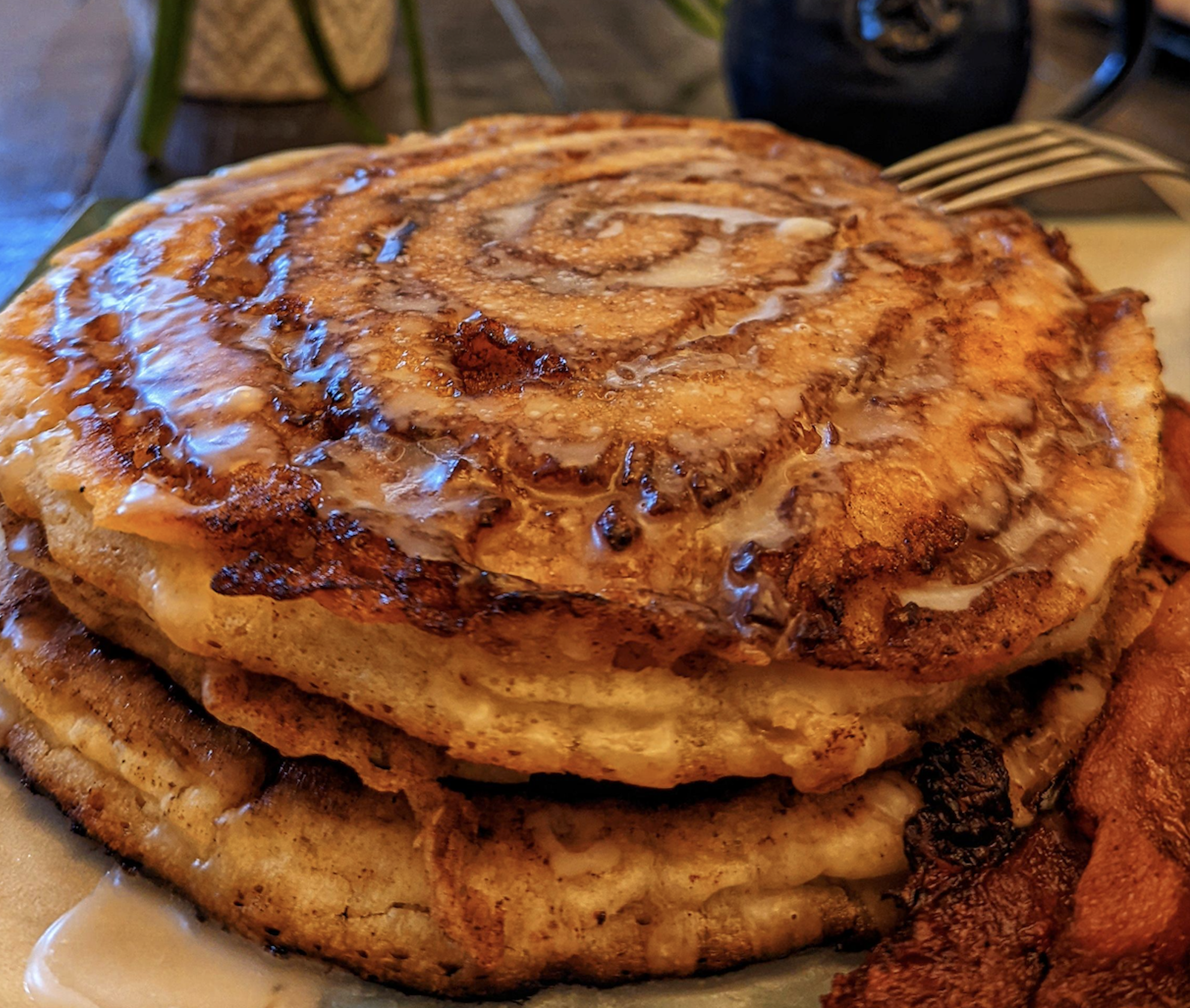 A stack of cinnamon roll pancakes drizzled with glaze, served with a side of bacon.