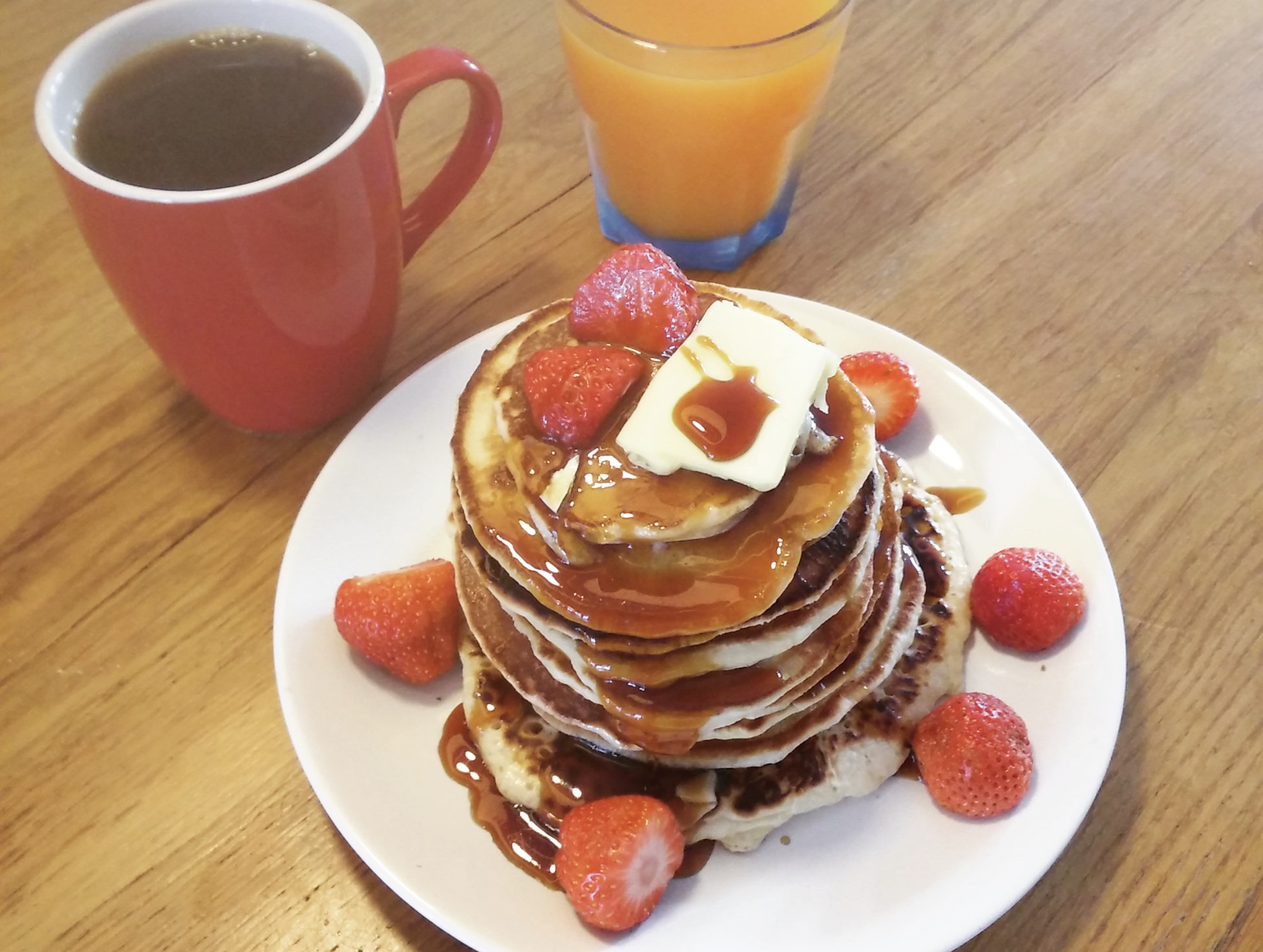 A stack of pancakes topped with butter, syrup, and strawberries on a plate, with a mug of coffee and a glass of orange juice nearby.