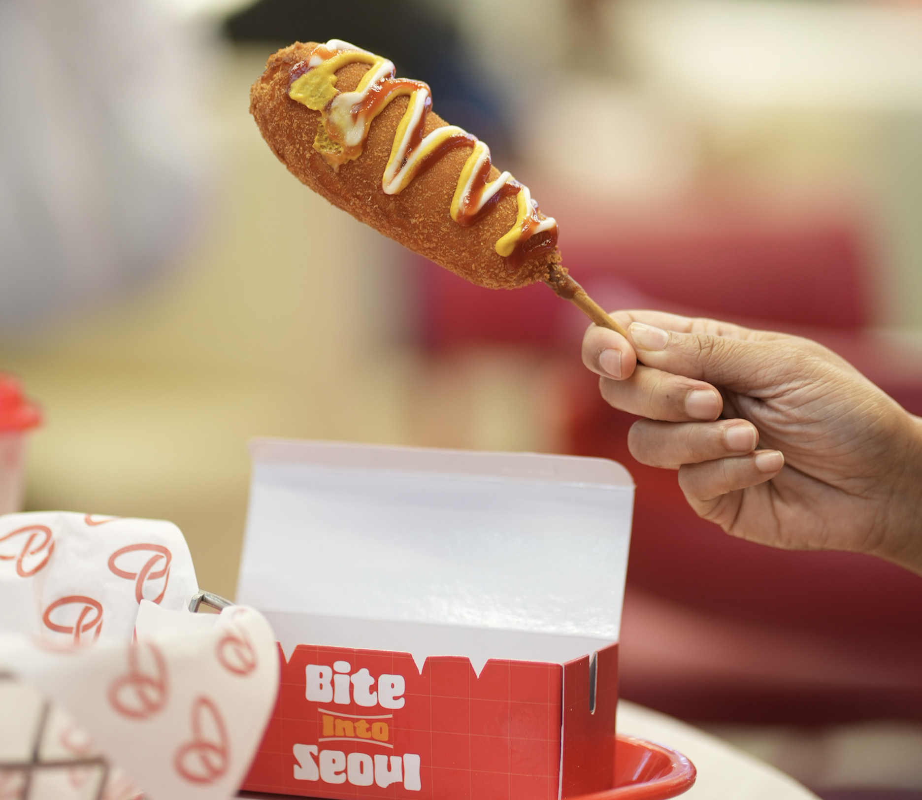 A hand holds a deep-fried corn dog drizzled with mustard and ketchup over a red takeout box labeled “Bite Into Seoul.”