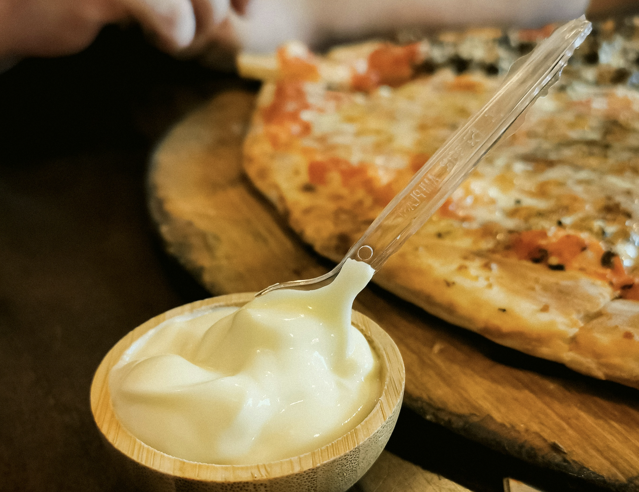 A small wooden bowl of white sauce with a clear plastic spoon, set in front of a pizza.