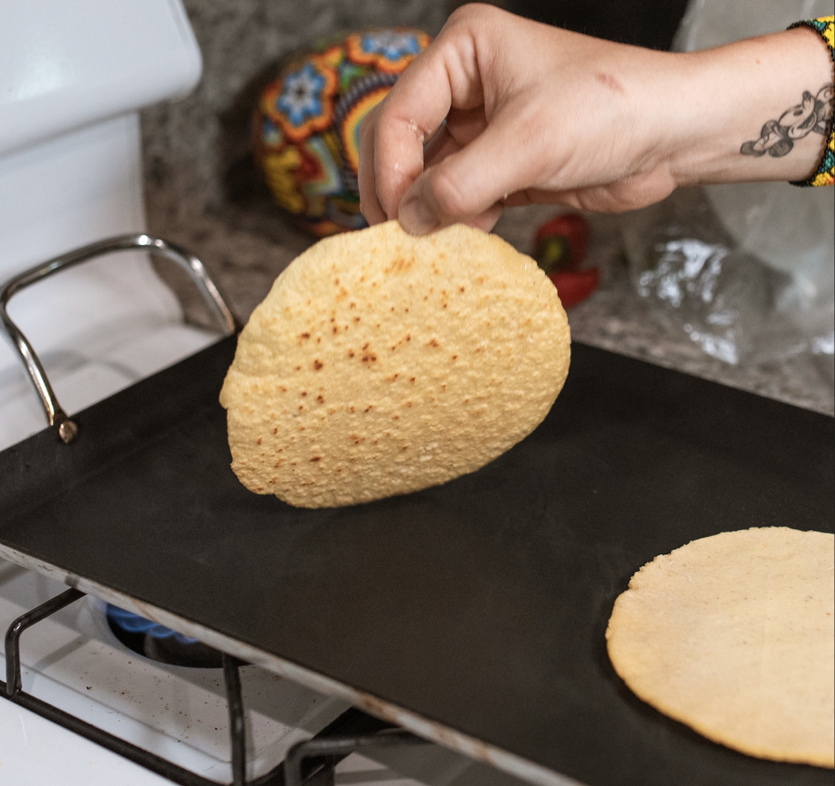 A hand places a corn tortilla onto a hot griddle.