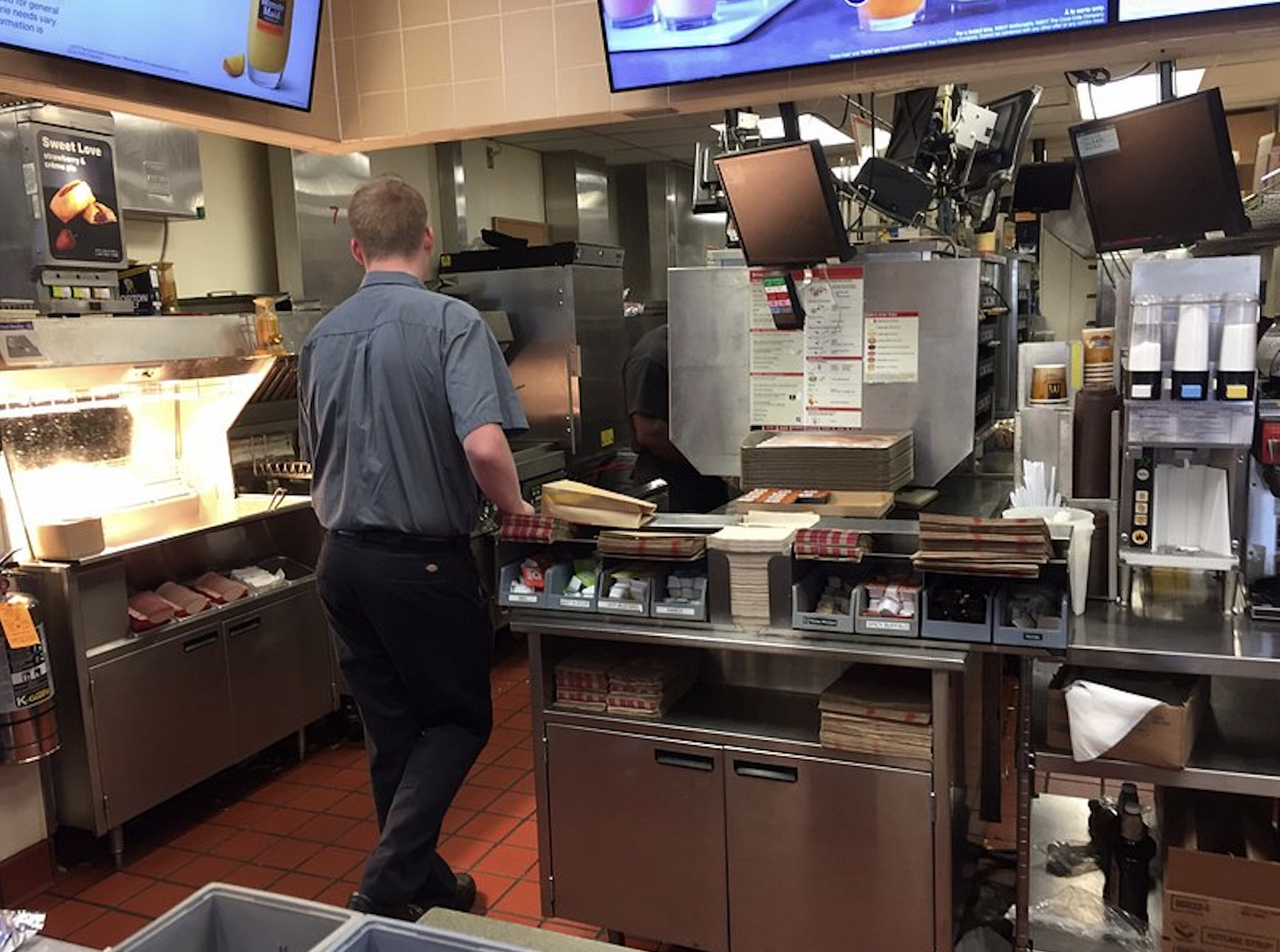 A restaurant employee in a gray uniform stands at a stainless steel prep station in a fast-food kitchen.