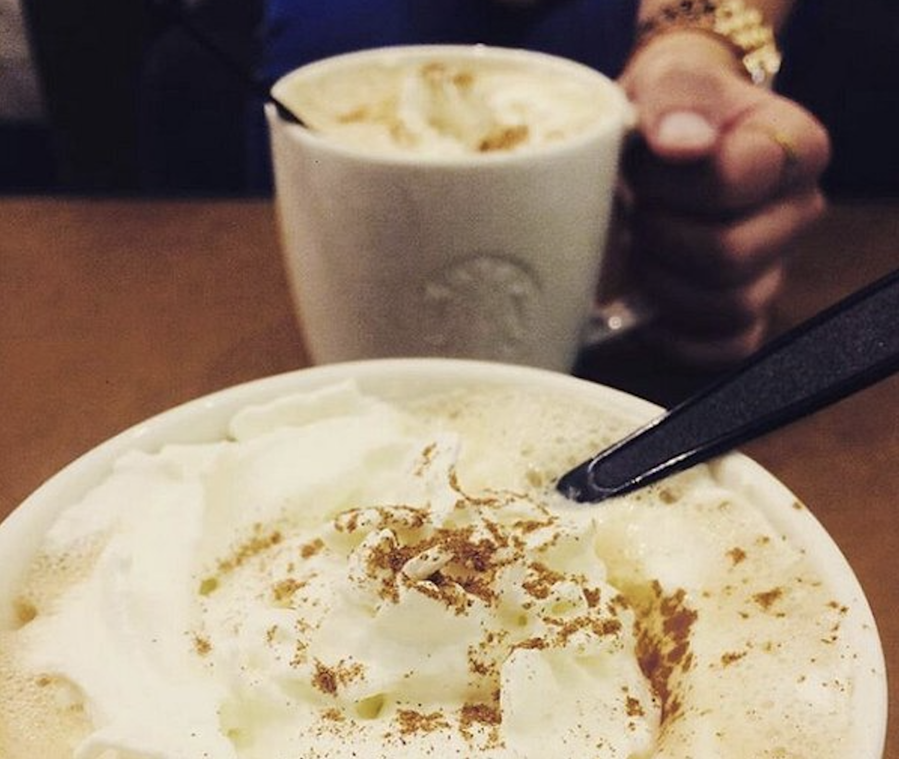 Two mugs of coffee topped with whipped cream and cinnamon, one held by a person in the background.