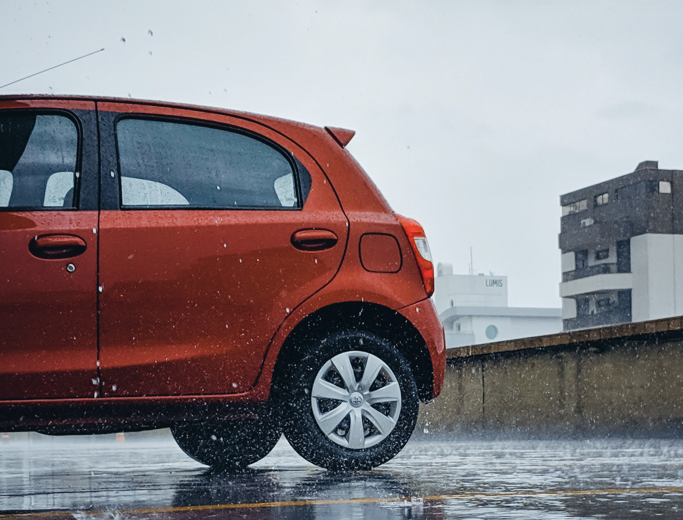 A side view of a red hatchback parked on wet pavement during a rain shower.