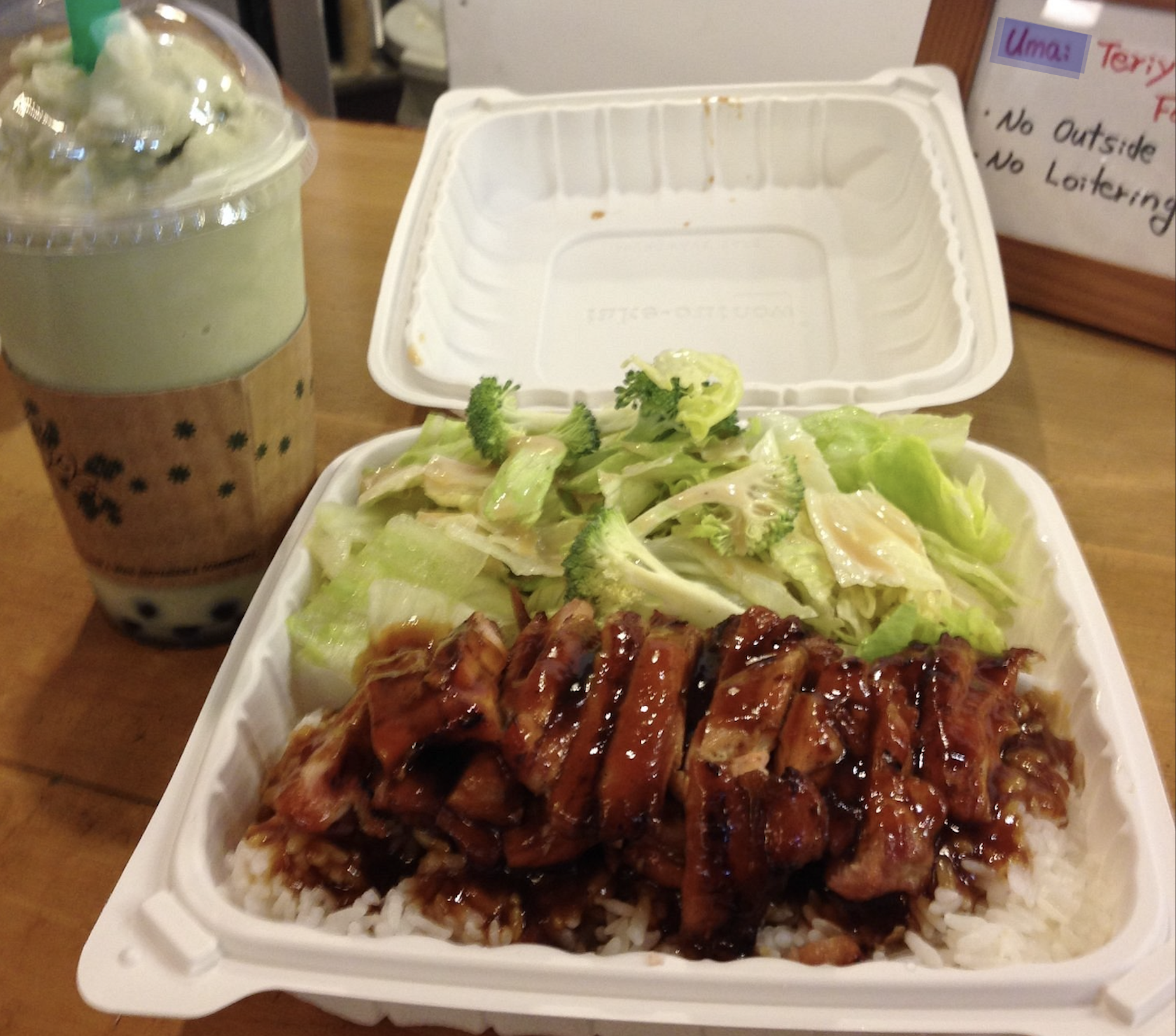 A takeout container with teriyaki chicken, rice, and a side salad, accompanied by a green boba tea.