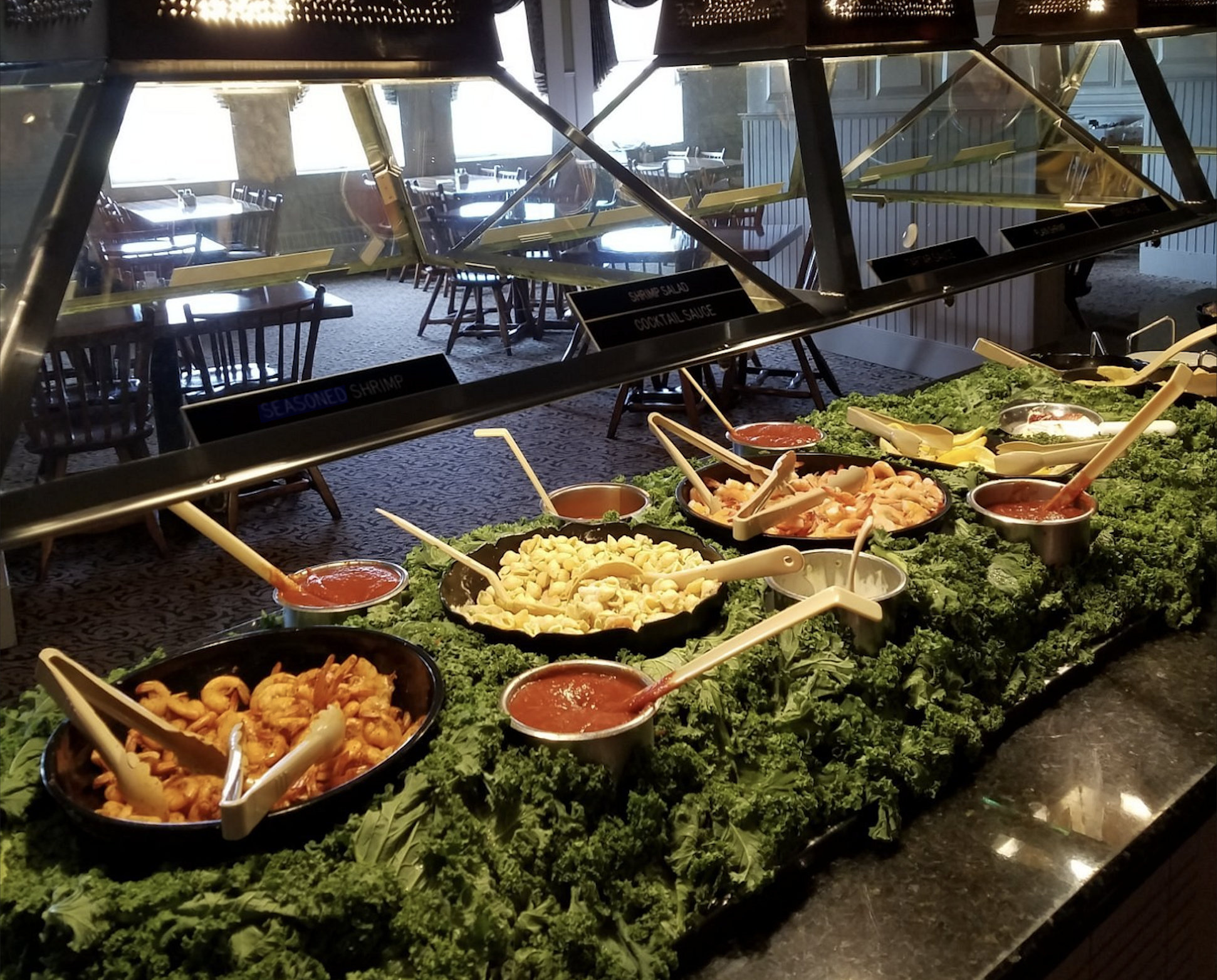 A buffet line featuring multiple bowls of shrimp, macaroni, and sauces arranged on a bed of fresh kale.