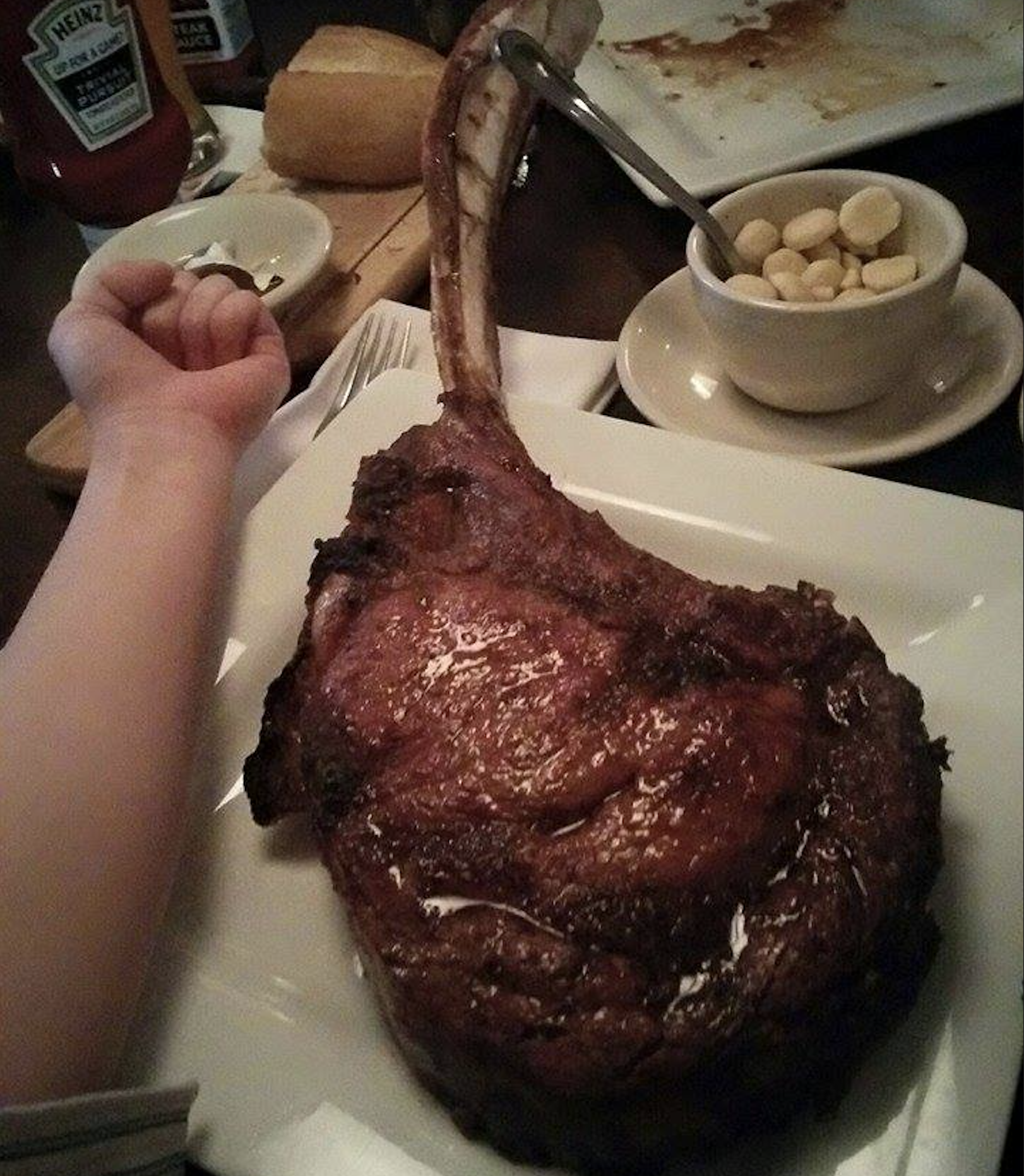 A large tomahawk steak on a white plate next to a person’s arm for scale.