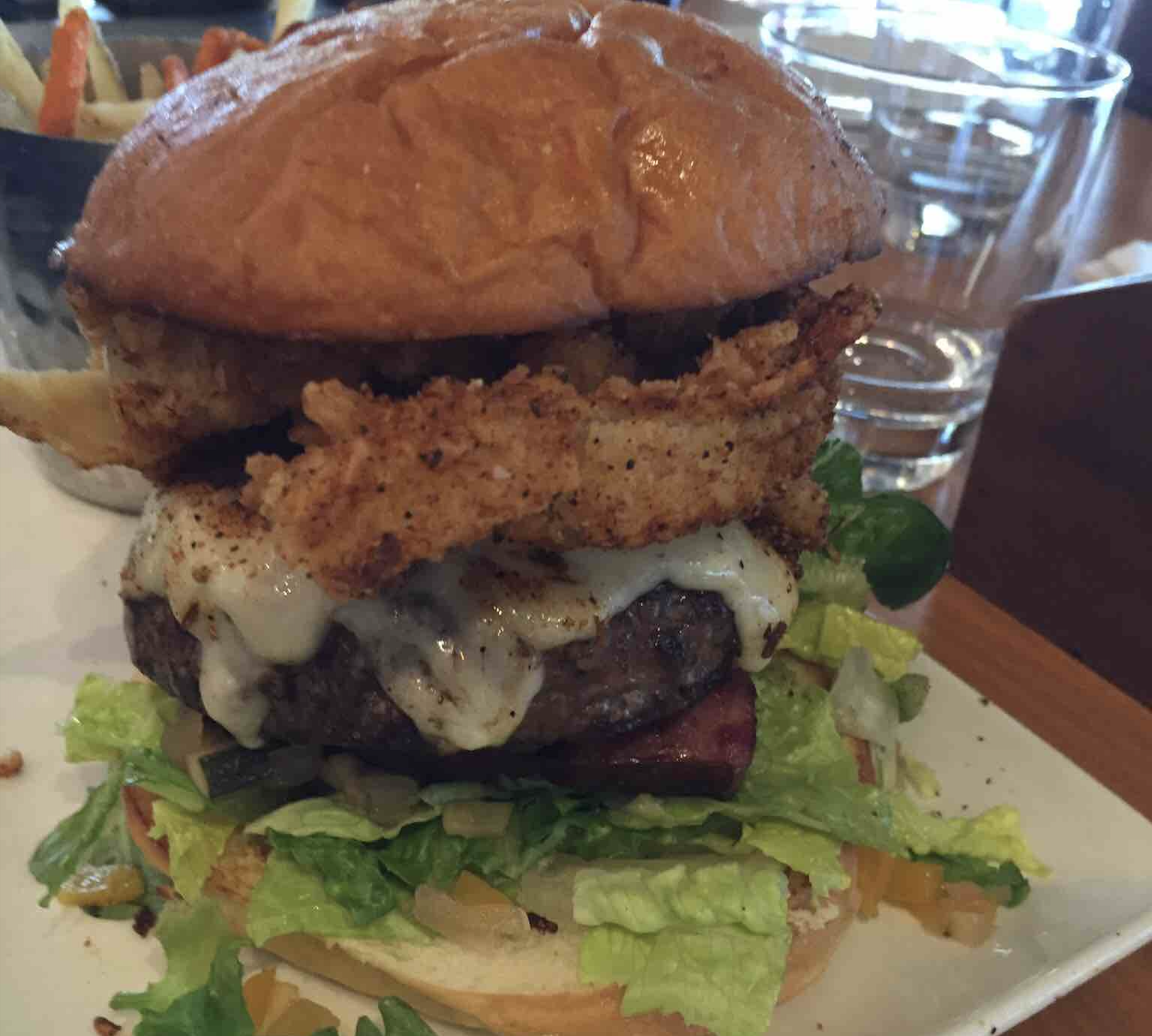 A burger topped with melted cheese and a large onion ring on a bed of lettuce, served on a plate with a side of fries.