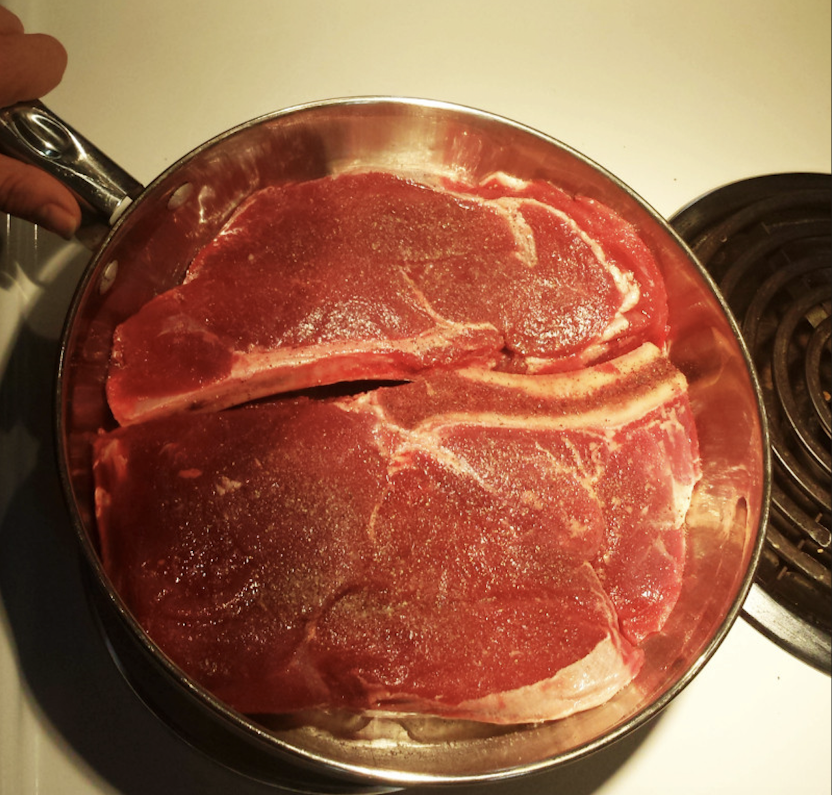 Two raw, seasoned steaks in a metal frying pan held over a stovetop.
