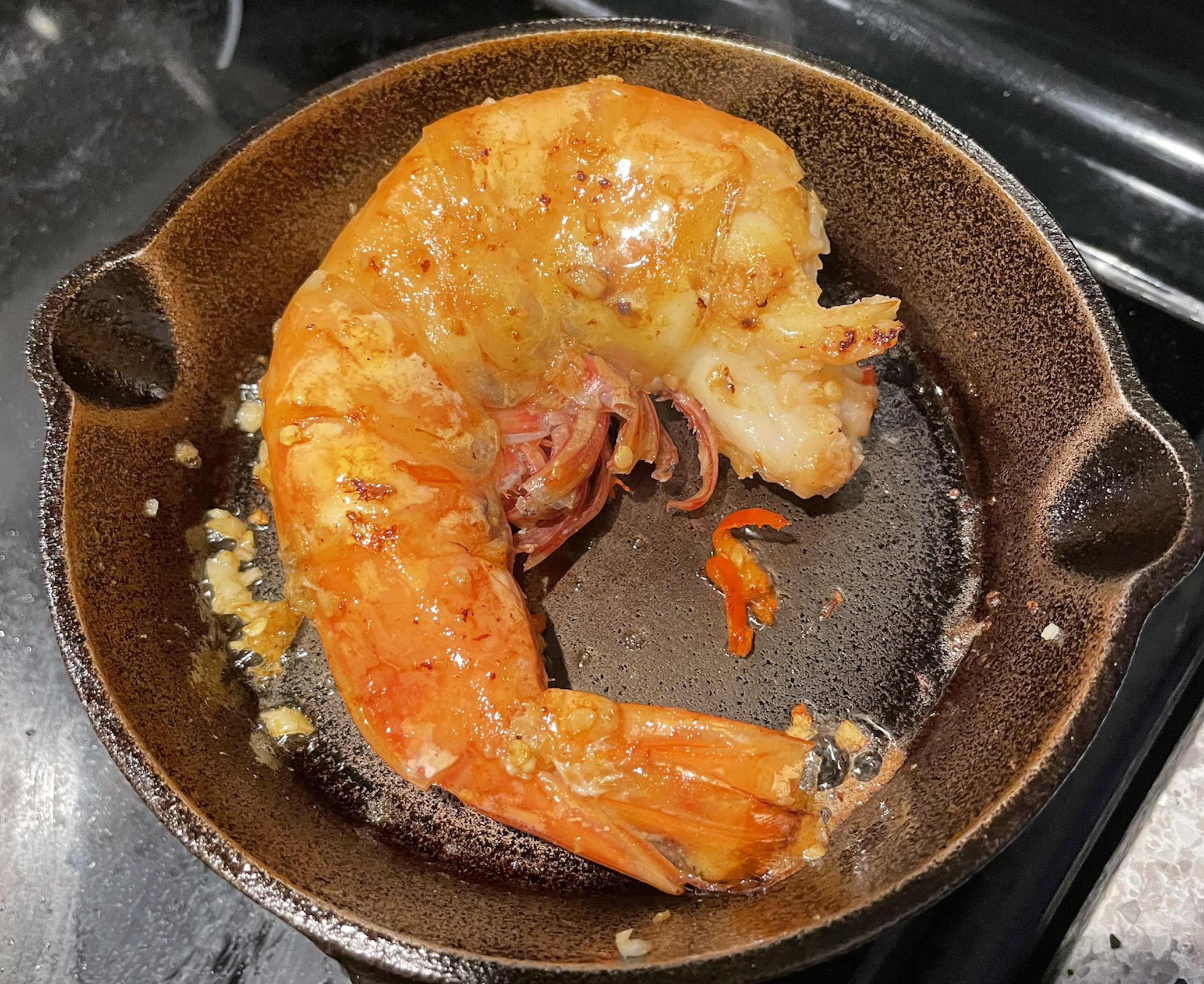 A single large, cooked shrimp with garlic and chili in a small cast-iron skillet.