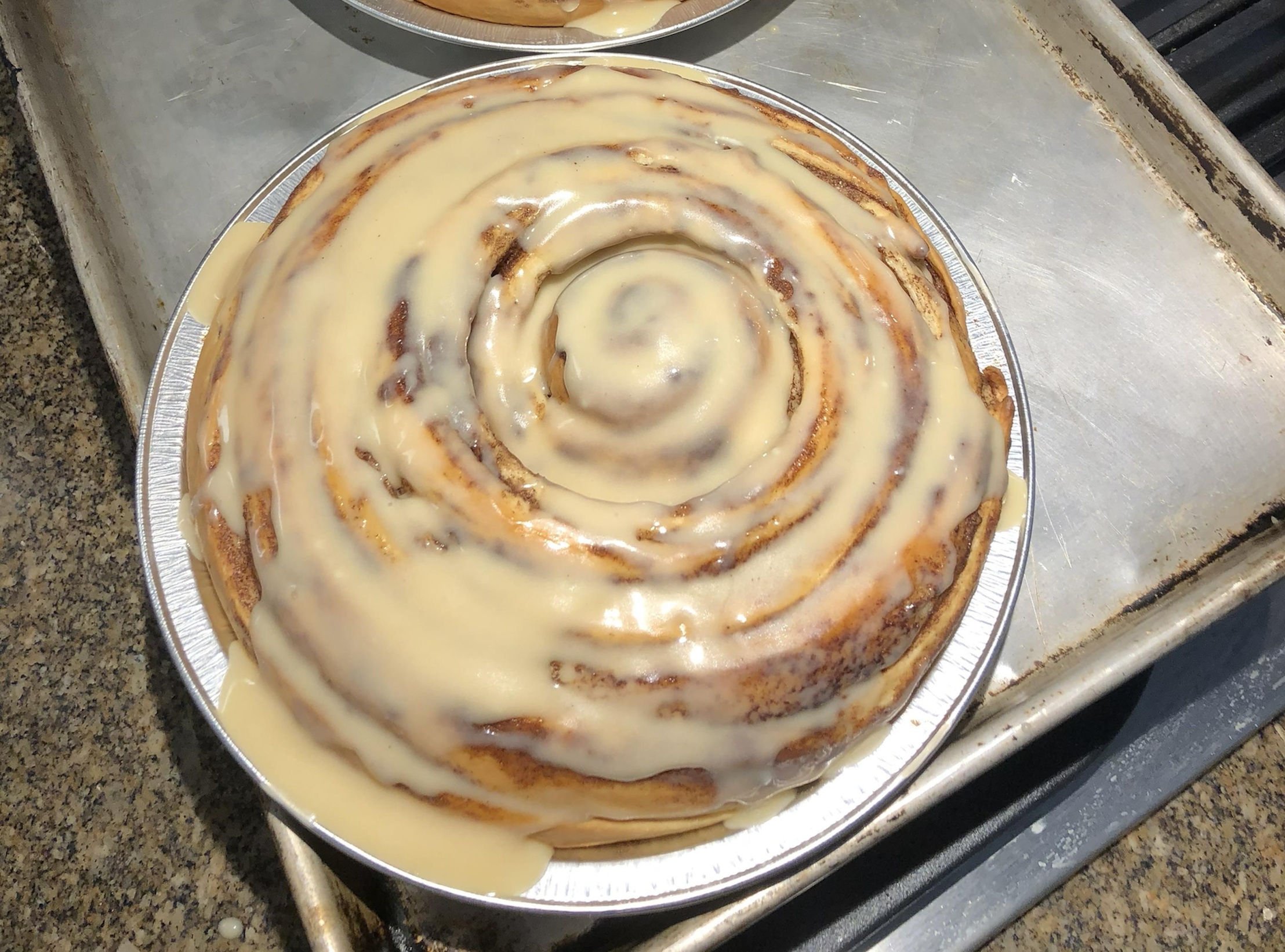 A large cinnamon roll covered in white icing in a round foil pan on a baking sheet.