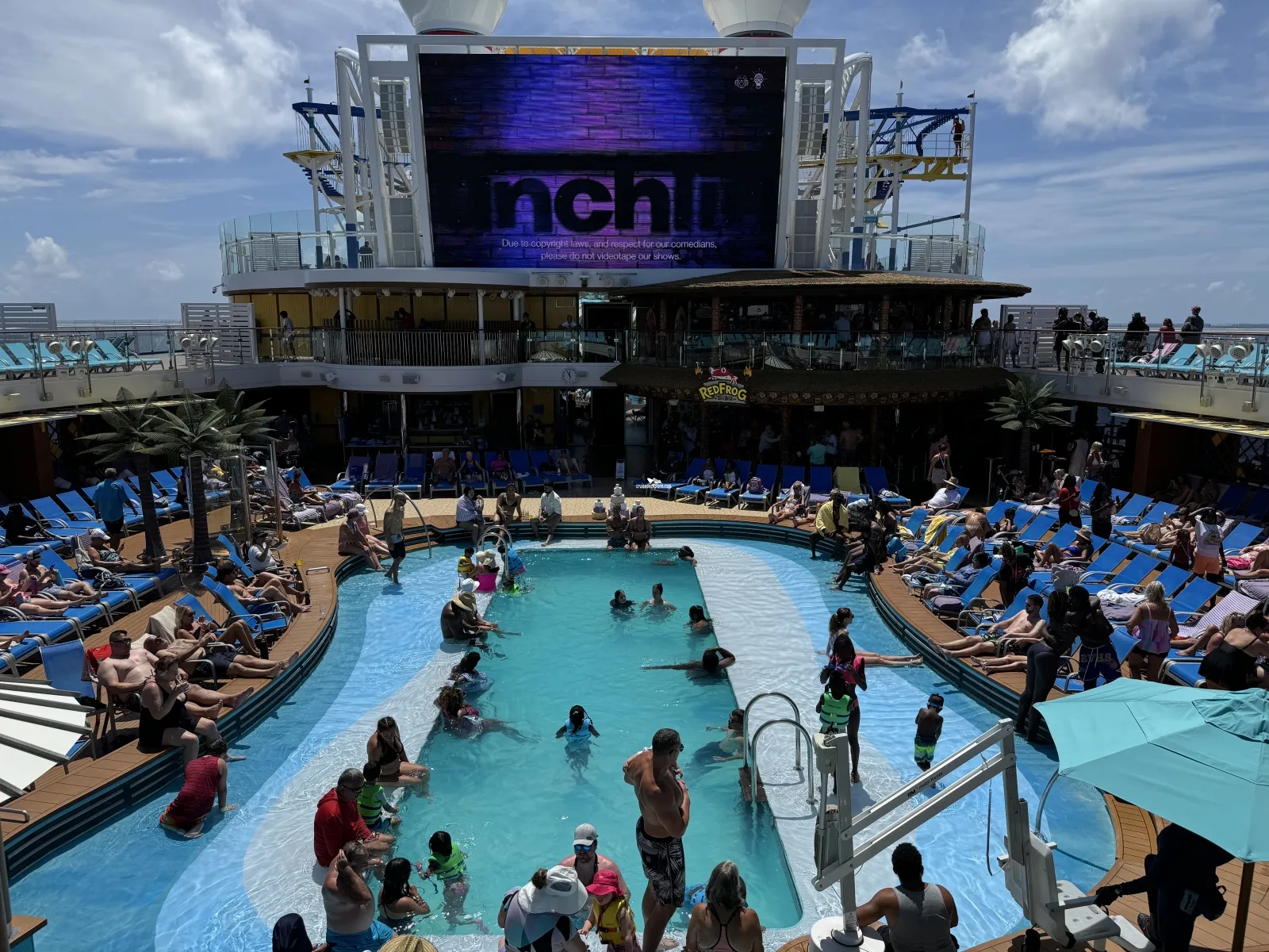 People swimming and relaxing in a cruise ship pool beneath a large outdoor screen displaying a notice about recording comedy shows.