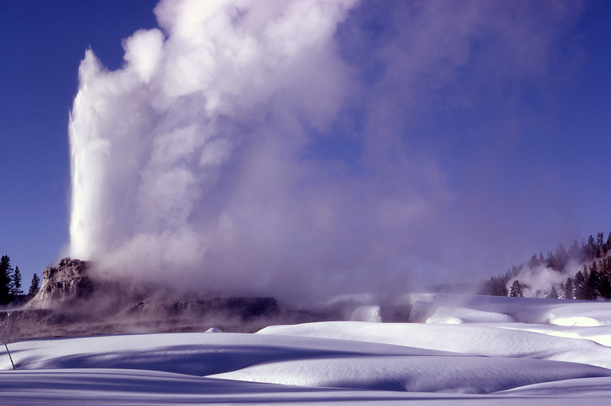 A geyser erupting steam against a blue sky, rising above a snow-covered landscape.