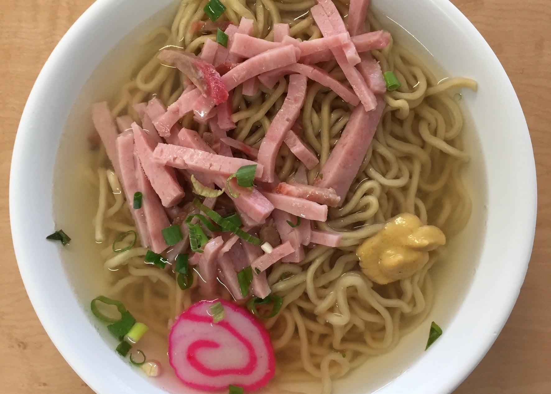 A bowl of ramen noodles in broth topped with strips of ham, a slice of pink-swirled fish cake, chopped green onions, and a dollop of mustard.