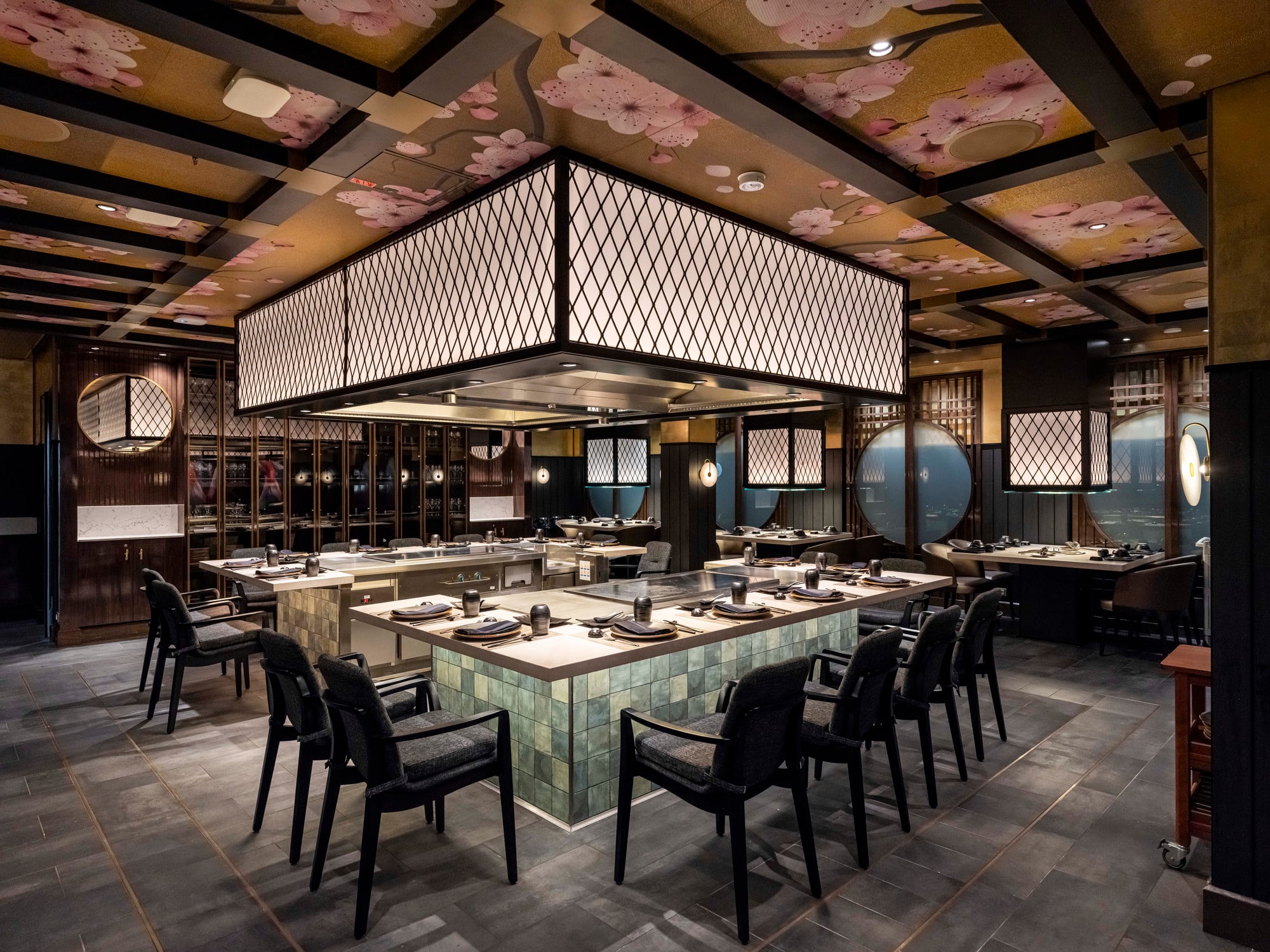 A Japanese-style teppanyaki restaurant interior featuring central cooking stations with square lantern-style light fixtures and a ceiling patterned with cherry blossoms.