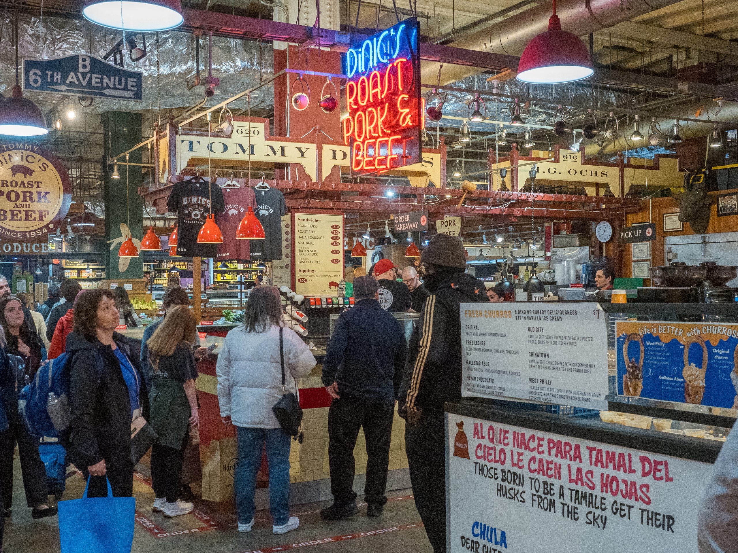 market stalls serving food People stand in line at Tommy DiNic’s roast pork and beef stand inside a food hall, with menu boards for sandwiches and churros visible.