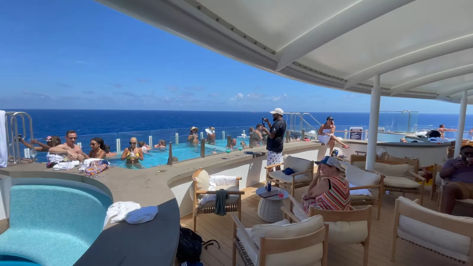 People relax in and around an infinity pool on the deck of a cruise ship overlooking the ocean.