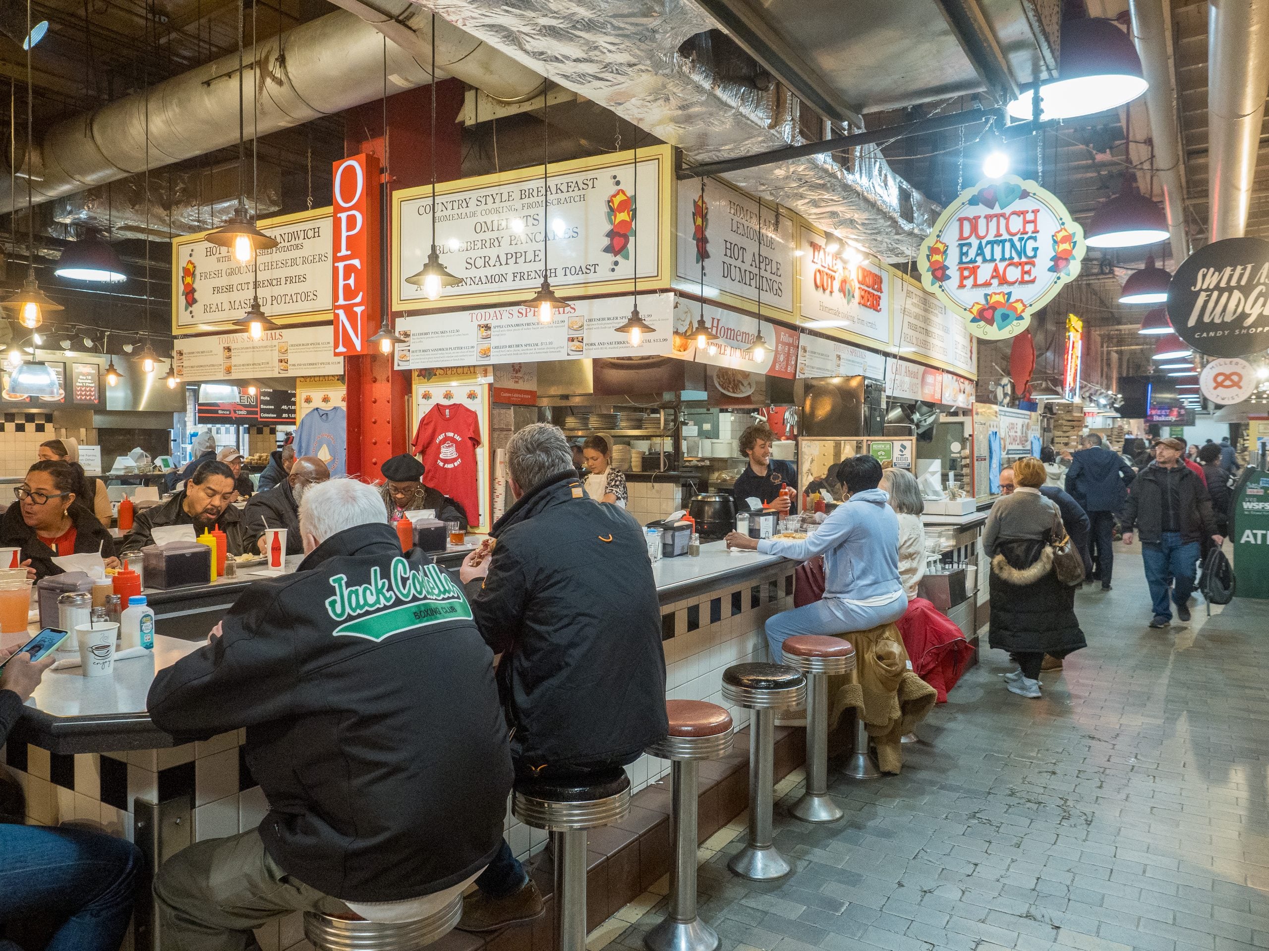 People sit at the counter of the Dutch Eating Place, one of the market stalls serving food inside an indoor food market.