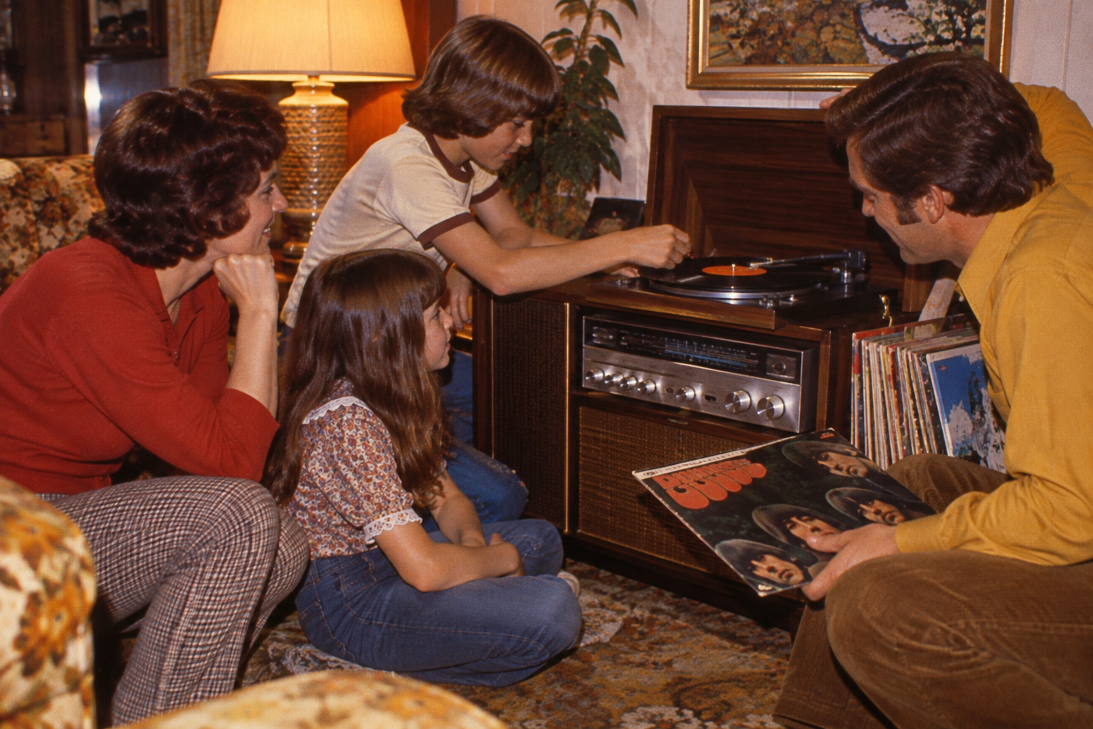 A family gathered around a vintage wooden console record player as a boy places a record on the turntable and a man holds a Beatles album cover.